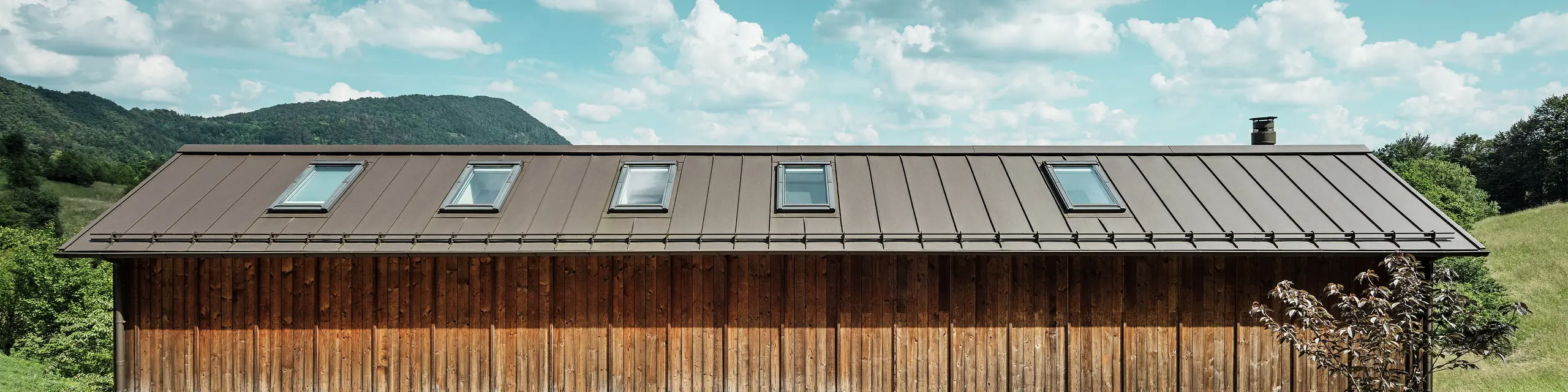 Cette maison moderne construite en pente séduit par sa façade en bois minimaliste et son toit en aluminium PREFALZ durable de couleur P.10 brun. La couverture à joint debout convainc par sa grande résistance aux intempéries et sa longévité, ainsi que par son harmonie avec l’environnement naturel. Des fenêtres de toit intégrées et des lignes épurées soulignent l’architecture intemporelle. Sur la photo, on distingue l’entrée de cette petite maison individuelle.