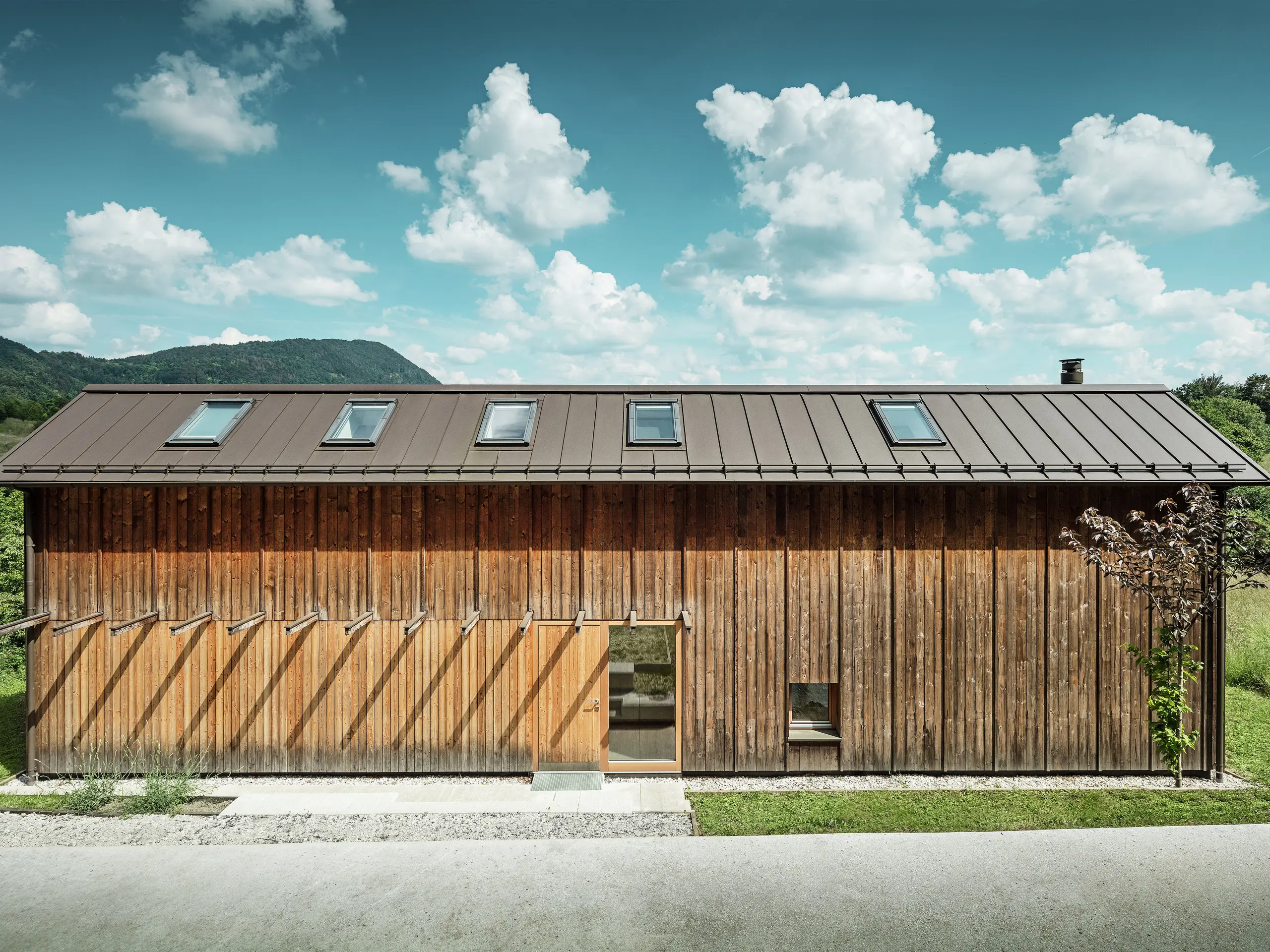 Cette maison moderne construite en pente séduit par sa façade en bois minimaliste et son toit en aluminium PREFALZ durable de couleur P.10 brun. La couverture à joint debout convainc par sa grande résistance aux intempéries et sa longévité, ainsi que par son harmonie avec l’environnement naturel. Des fenêtres de toit intégrées et des lignes épurées soulignent l’architecture intemporelle. Sur la photo, on distingue l’entrée de cette petite maison individuelle.