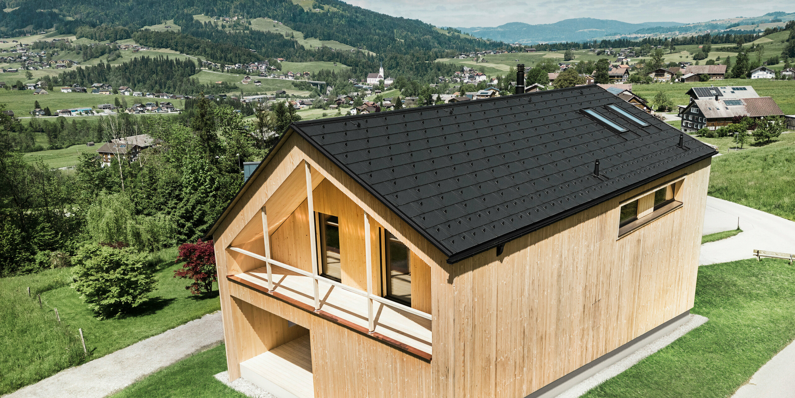 Cette photo montre une maison vitrine écologique à Egg, qui se distingue par sa tuile solaire PREFA et ses R.16 noirs dans le décor pittoresque d'un village autrichien. Les éléments solaires sont parfaitement intégrés dans la structure du toit et fournissent de l'énergie à partir d'une source d'énergie renouvelable, tandis que le bois naturel de la façade et l'environnement verdoyant représentent une combinaison de durabilité et de localité. La vue sur la structure du village environnant avec les bâtiments traditionnels en arrière-plan donne à l'image une dimension supplémentaire et souligne l'intégration réussie de méthodes de construction modernes et respectueuses de l'environnement dans les paysages établis.