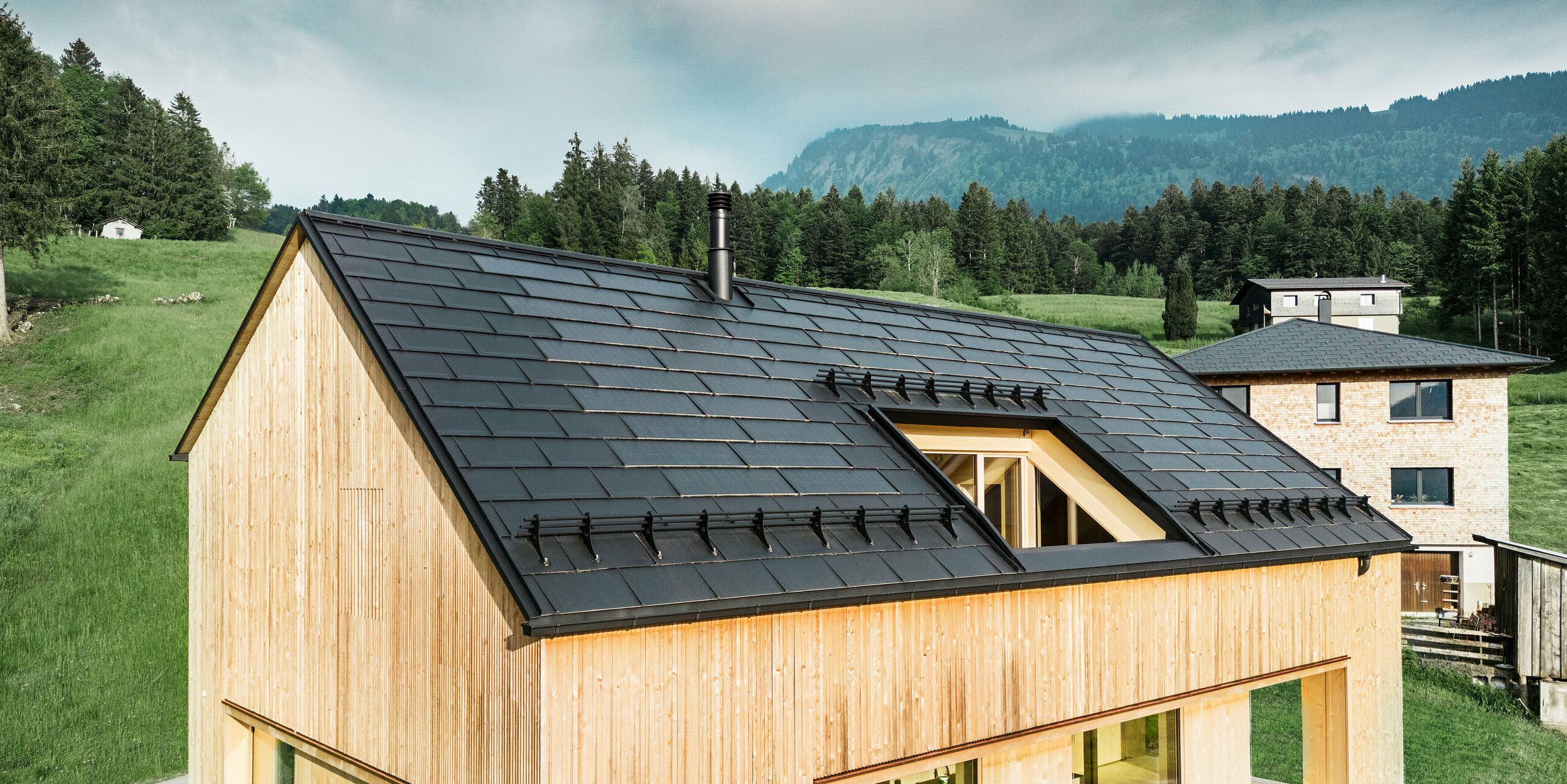 Vorderansicht eines modernen Holzhauses in Egg, das sich mit seinem dunklen PREFA Solardach vor der üppigen, grünen Landschaft abzeichnet. Die präzise verlegten schwarzen PREFA Solardachplatten bieten nicht nur Schutz, sondern auch Energieeffizienz durch die integrierte Solartechnologie. An den Rändern des Daches bis hin zur Traufe wurden traditionelle PREFA Dachplatten R.16 in schwarz verlegt. Der klassische Schornstein und das große Dachfenster ergänzen das nachhaltige Konzept des Gebäudes, das moderne Architektur und umweltbewusste Baupraktiken in Einklang bringt.