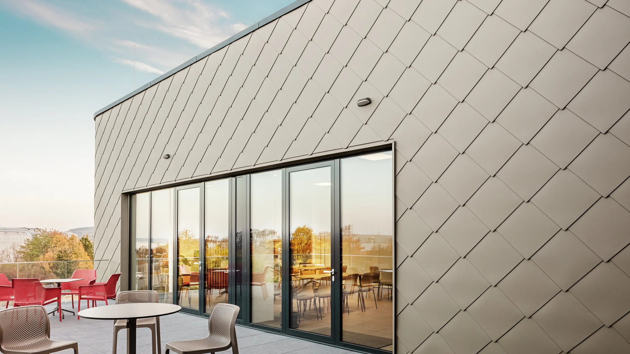 Die Fassade des neuen PREFA Standortes in Törökbálint besticht durch die eleganten PREFA Wandrauten 44 × 44 in P.10 Prefabronze. Die präzise verlegte Aluminiumfassade sorgt für ein modernes, dynamisches Erscheinungsbild und bietet gleichzeitig maximalen Schutz vor Witterungseinflüssen. Die großzügige Glasfront verbindet Innen- und Außenbereich und schafft auf der großzügigen Terrasse eine helle und einladende Atmosphäre.