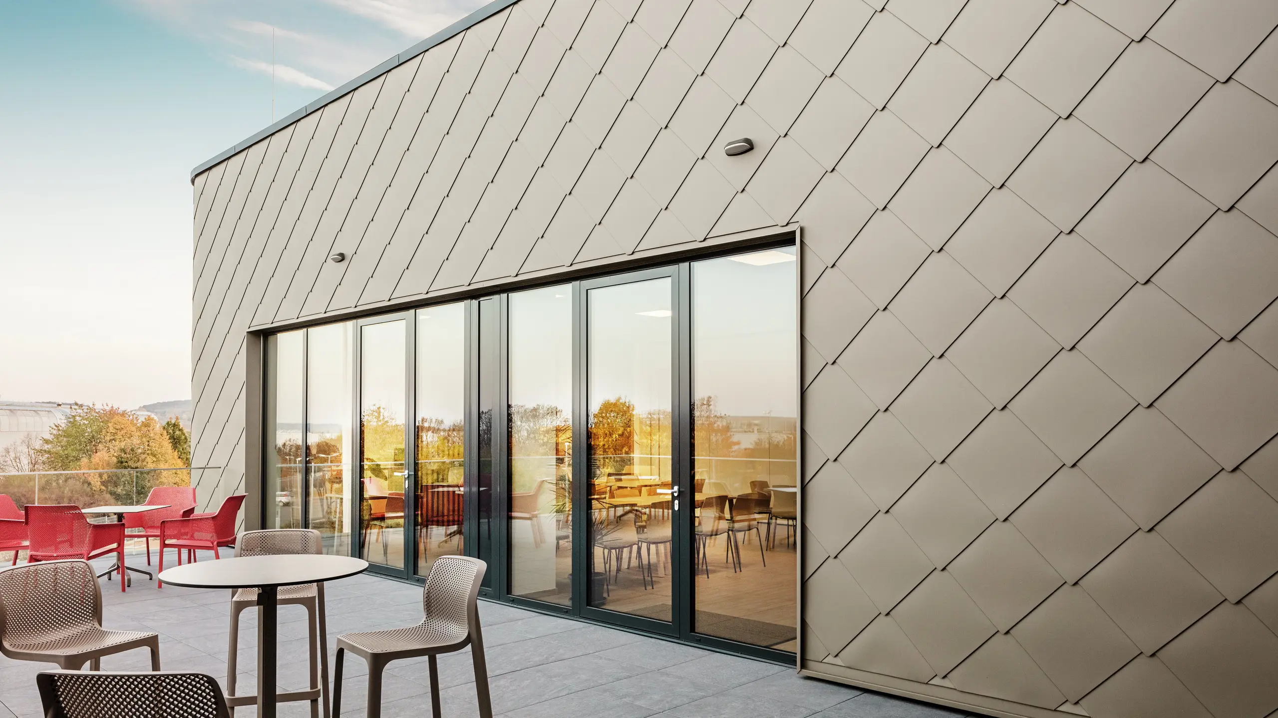 La façade du nouveau site PREFA à Törökbálint impressionne par les élégants losanges de façade PREFA 44 × 44 en P.10 bronze prefa. La façade en aluminium posée avec précision crée une apparence moderne et dynamique tout en offrant une protection maximale contre les éléments. La généreuse façade vitrée relie les espaces intérieurs et extérieurs et crée une atmosphère lumineuse et accueillante sur la terrasse spacieuse.