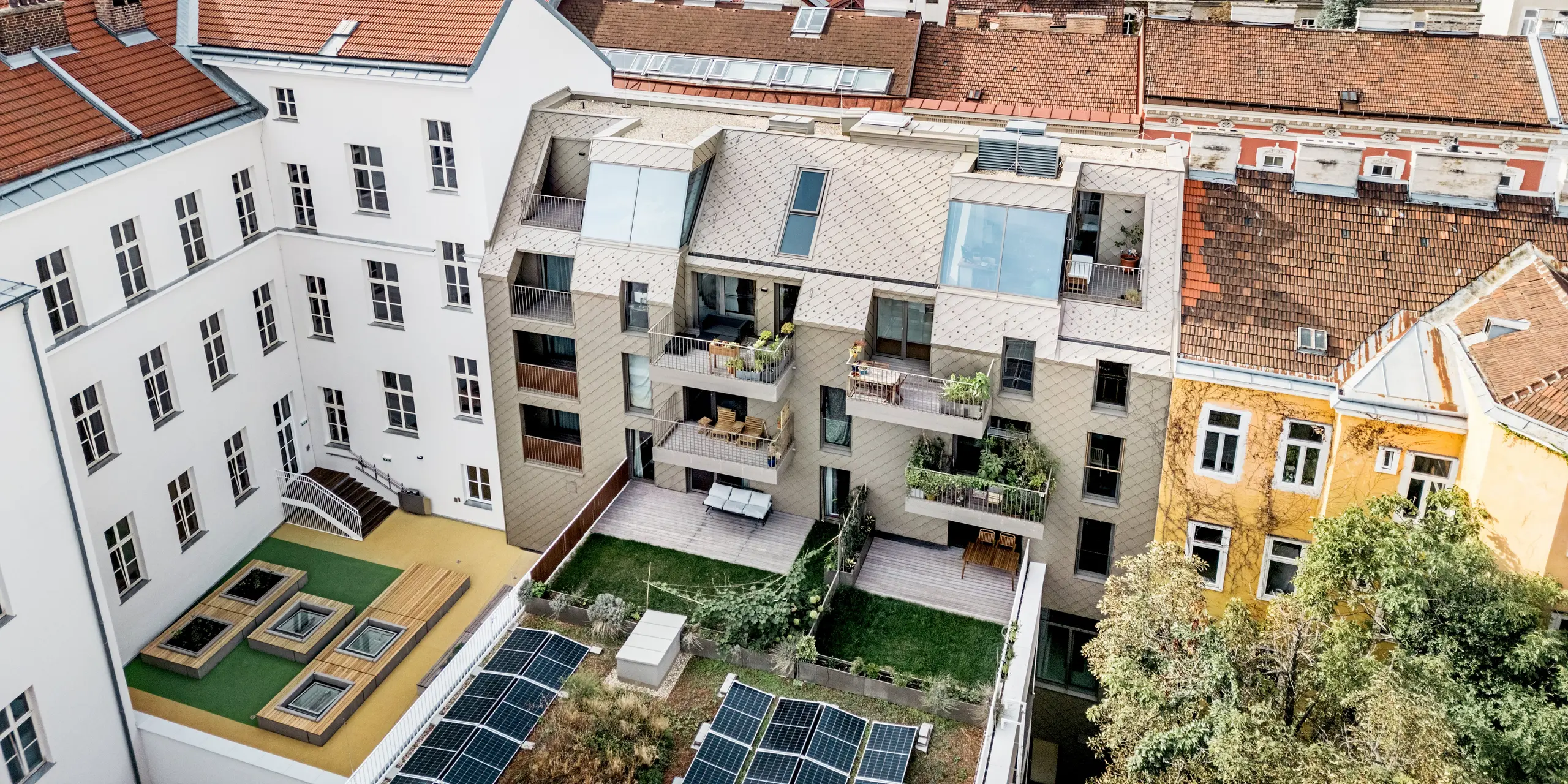 La vue aérienne révèle un panorama exceptionnel de l'extension d'un immeuble résidentiel à Vienne, où des losanges 29x29 dans la teinte bronze ont été utilisés sur le toit et la façade. Ces losanges modernes s'harmonisent parfaitement avec l'architecture traditionnelle du quartier, créant un lien visuel attrayant entre ancien et nouveau. La cour intérieure, conçue avec soin, intègre des panneaux solaires, des espaces verts et des terrasses en bois, soulignant ainsi l'engagement du complexe résidentiel en faveur du développement durable. Le mariage harmonieux entre espace urbain et nature est sublimé par les terrasses et balcons sur le toit, qui offrent aux résidents des vues imprenables sur les toits viennois.