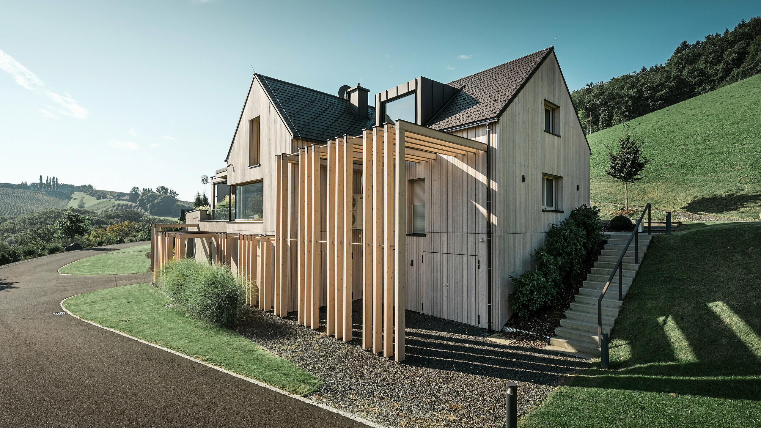Großes Einfamilienhaus mit Satteldach und Gaube in Hanglage. Das Dach ist mit braunen PREFA Aluminium Dachschindeln eingedeckt und die Fassade mit vertikalen Holzbrettern verkleidet.