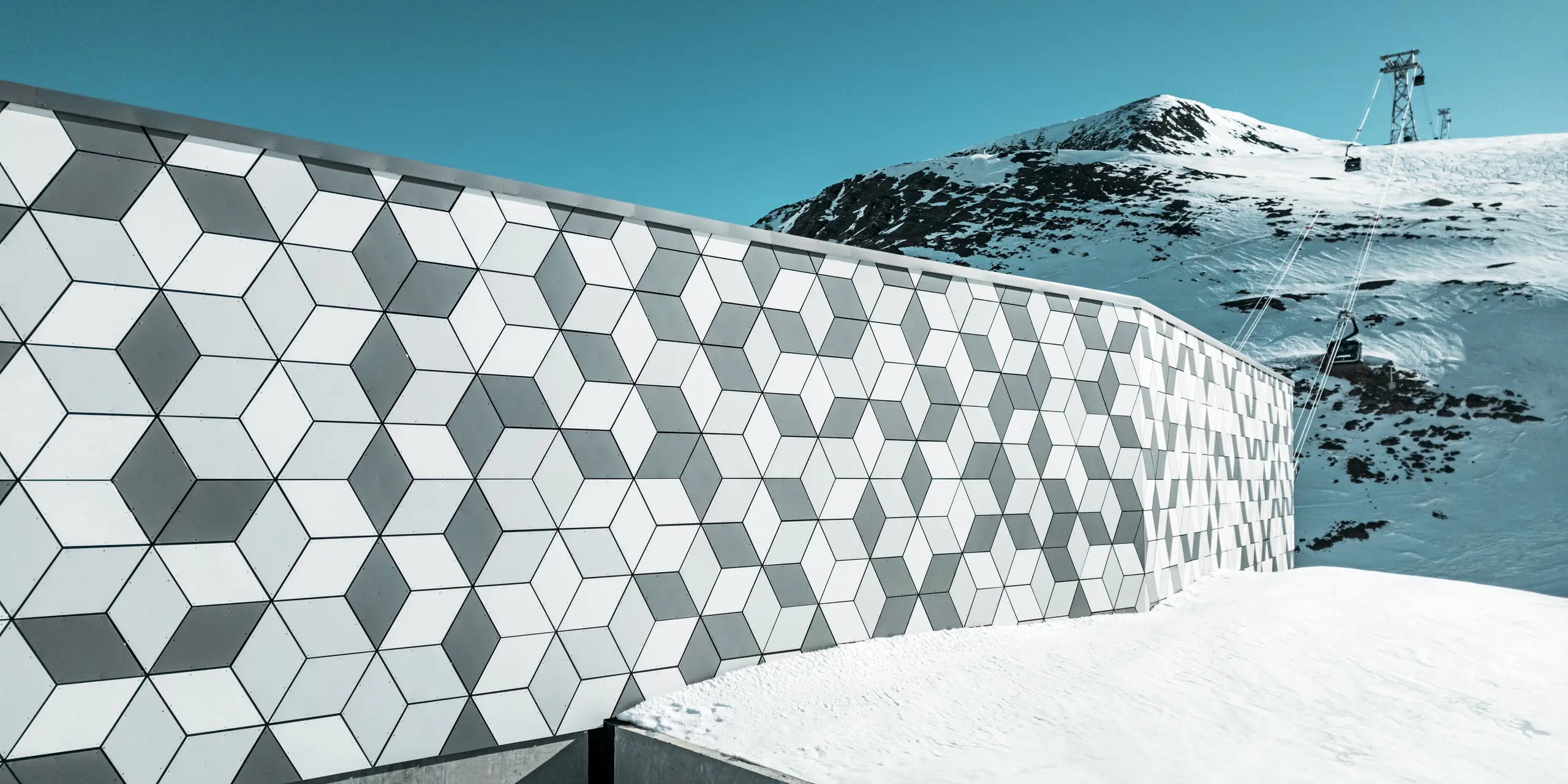 La façade de la station de téléphérique des Deux Alpes est un chef-d'œuvre d'architecture moderne. Elle est construite en panneaux composites en aluminium PREFABOND disposés selon un motif saisissant de losanges blancs et gris. Son design géométrique s'intègre harmonieusement au paysage montagneux enneigé et, grâce à des matériaux robustes et résistants aux intempéries, offre une protection durable contre les intempéries. La vue depuis la façade permet d'admirer le chemin de la télécabine qui monte vers la montagne.