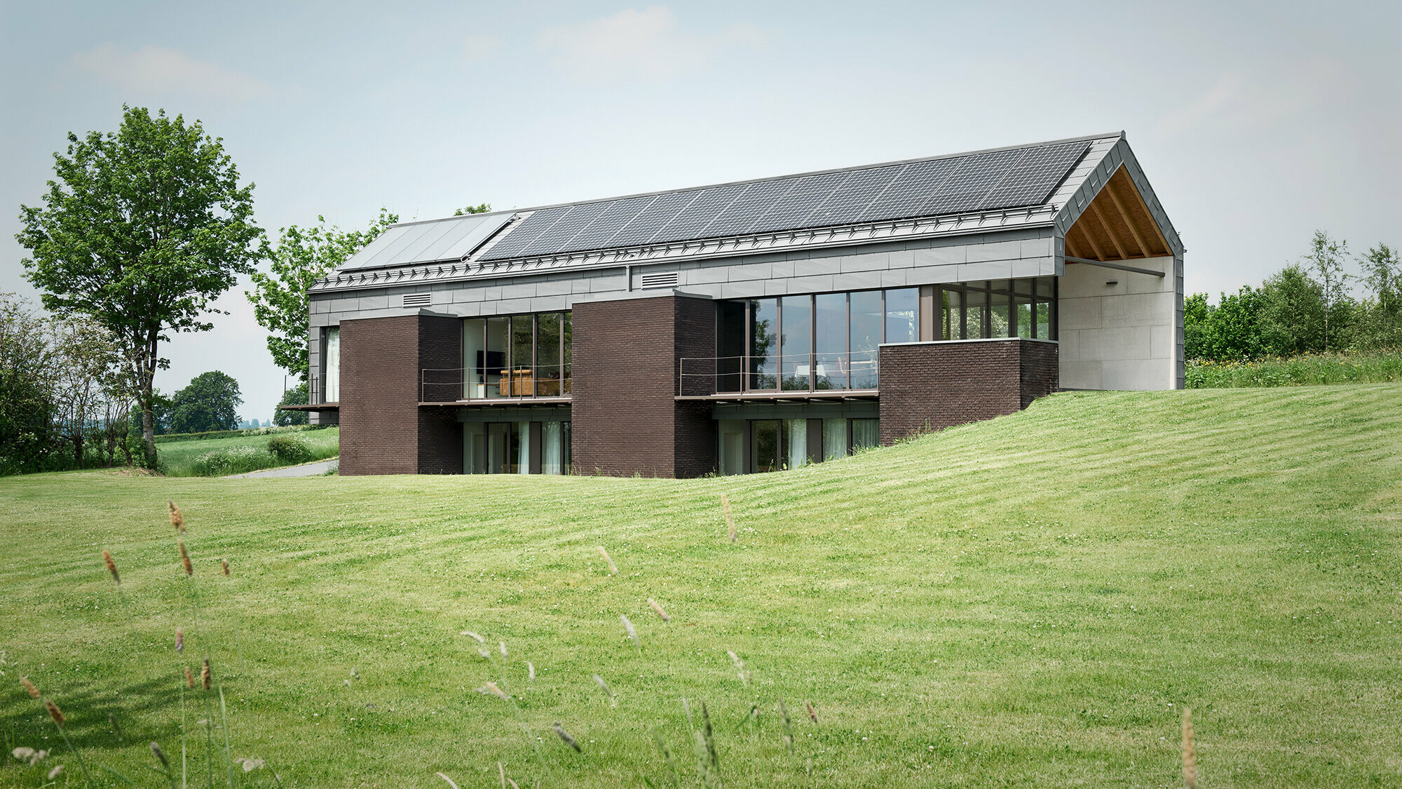 Blick auf den langgezogenen Baukörper von der südlichen Gartenseite und das harmonische Zusammenspiel von Backstein und den Aluminiumpaneelen von PREFA.