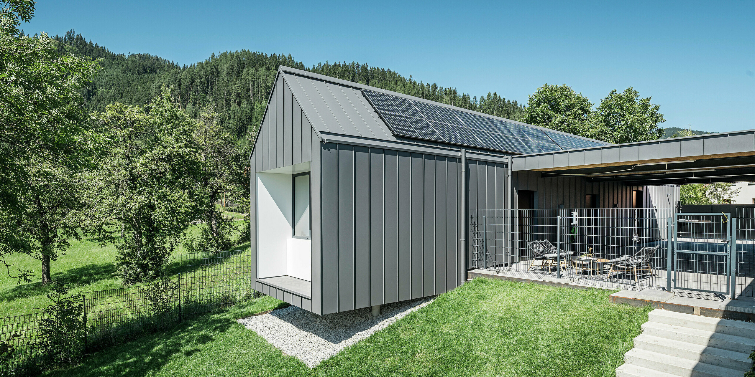 Harmonický zadní pohled na moderní rodinný dům v rakouském Pogier se střešním a fasádním systémem PREFALZ v tmavě šedé barvě P.10, integrovaným do idylické lesní krajiny. Kombinace tmavě šedého hliníkového opláštění a přírodního prostředí podtrhuje ekologický koncept domu doplněný o efektivní fotovoltaický systém na střeše. Oplocený venkovní areál s relaxačním posezením zve k lenošení a vychutnávání klidu a svěžesti okolního lesa.