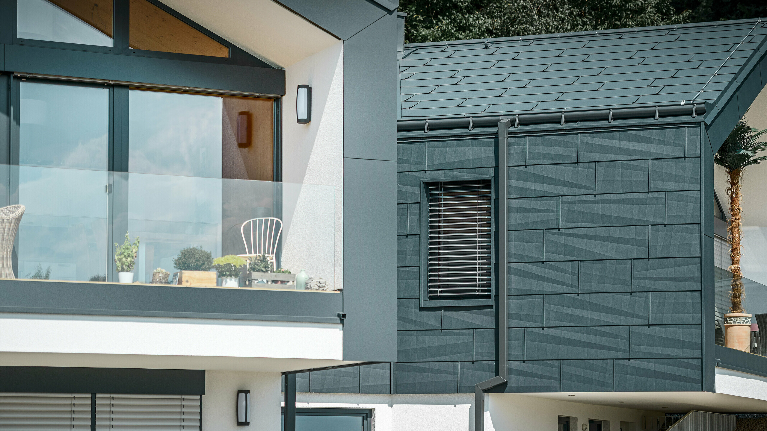 La photo montre une maison jumelée moderne avec une façade élégante en aluminium. La façade est peinte dans un ton anthracite foncé et présente une finition structurée grâce au panneau de toiture et de façade PREFA FX.12. Une gouttière havraise et un tuyau de descente carré PREFA ont été installés sur le toit. Un balcon avec une balustrade en verre est visible à l'étage supérieur, tandis que l'étage inférieur est doté de fenêtres avec des stores à lamelles. L'architecture est claire et minimaliste, et des arbres sont visibles en arrière-plan, créant une atmosphère naturelle.