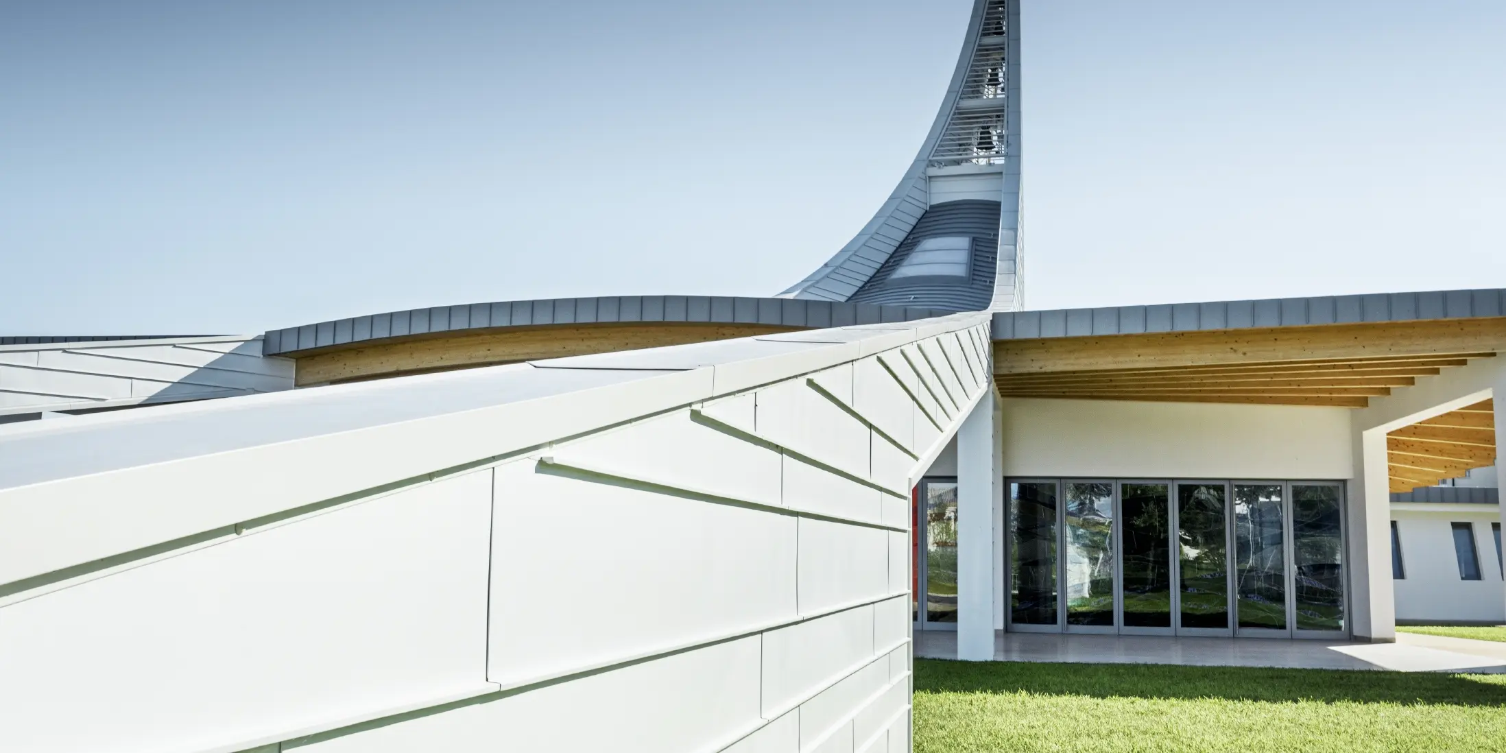 Modernes Kirchengebäude mit geschwungener Dachkonstruktion aus hellen Aluminiumpaneelen. Im Hintergrund erhebt sich ein hoher, elegant gebogener Turm mit einer Statue an der Spitze. Die Architektur kombiniert klare Linien mit offenen Glasflächen und Holzelementen unter dem Dach. Der Himmel ist wolkenlos und betont die helle, futuristische Gestaltung des Bauwerks