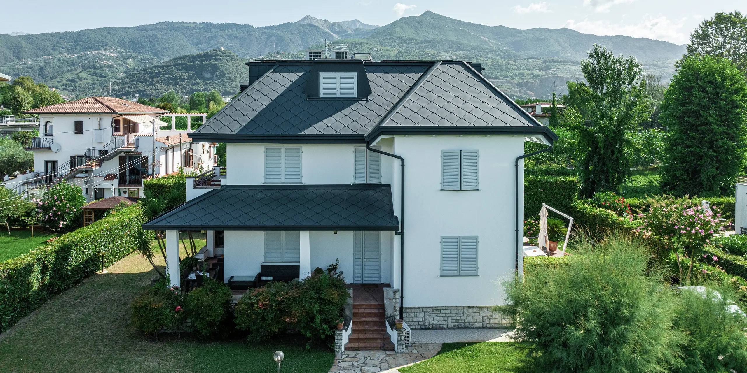 Vue de face d'une simple maison individuelle à Montignoso, Italie. Le toit avec le losange PREFA 44 × 44 en P.10 gris pierre se démarque parfaitement sur le ciel bleu vif. La couverture en aluminium de haute qualité assure un look moderne et élégant tout en offrant une protection maximale contre les conditions météorologiques extrêmes. La combinaison de la façade blanche, du toit sombre et de l'environnement vert crée une image globale harmonieuse.