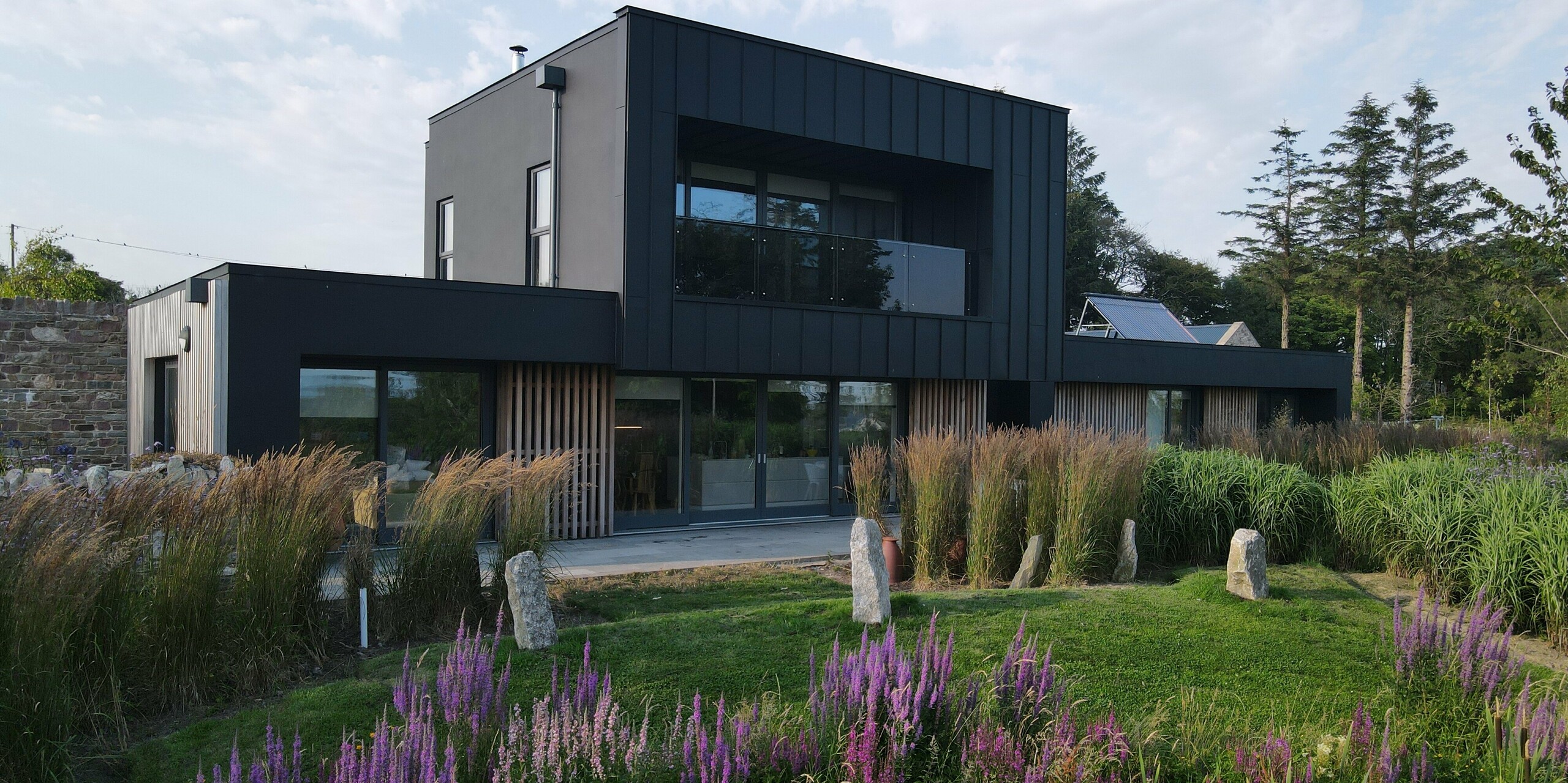 Das Bild zeigt das moderne Architektenhaus in Wexford, das von einer üppigen, naturnahen Gartenlandschaft mit hohen Gräsern und violett blühendem Lavendel umgeben ist. Die schwarze PREFALZ-Fassade in der Farbe P.10 Schwarz prägt das markante Erscheinungsbild des Gebäudes mit ihren klaren Linien, dem tiefen Matt-Schwarz und der präzisen vertikalen Struktur. Die großzügigen Fensterfronten und der überdachte Balkon unterstreichen den modernen Charakter. Die robuste PREFA-Aluminiumfassade ist witterungsbeständig und langlebig. Sie bildet einen spannenden Kontrast zur lebendigen Vegetation im Vordergrund.