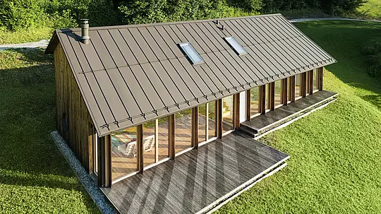 Maison en bois moderne avec une grande façade vitrée et un toit en aluminium PREFALZ résistant de couleur P.10 brun – idéale pour les terrains en pente comme à Vinje. La couverture de toit de haute qualité offre une résistance maximale aux intempéries, une longue durée de vie et une esthétique élégante. Grâce à la pose précise des joints debout et à l’intégration de fenêtres de toit, un concept d’habitat durable et baigné de lumière voit le jour. La vue en plongée met en évidence la finition soignée des joints debout en aluminium.