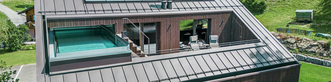 Ansicht eines modernen Mehrfamilienhauses mit einem PREFA Dach in Prefalz-Technik, ausgeführt in der Farbe Nussbraun. Das Bild zeigt auch eine Terrasse mit einem Pool, umgeben von einer Holzverkleidung und Glasgeländern.