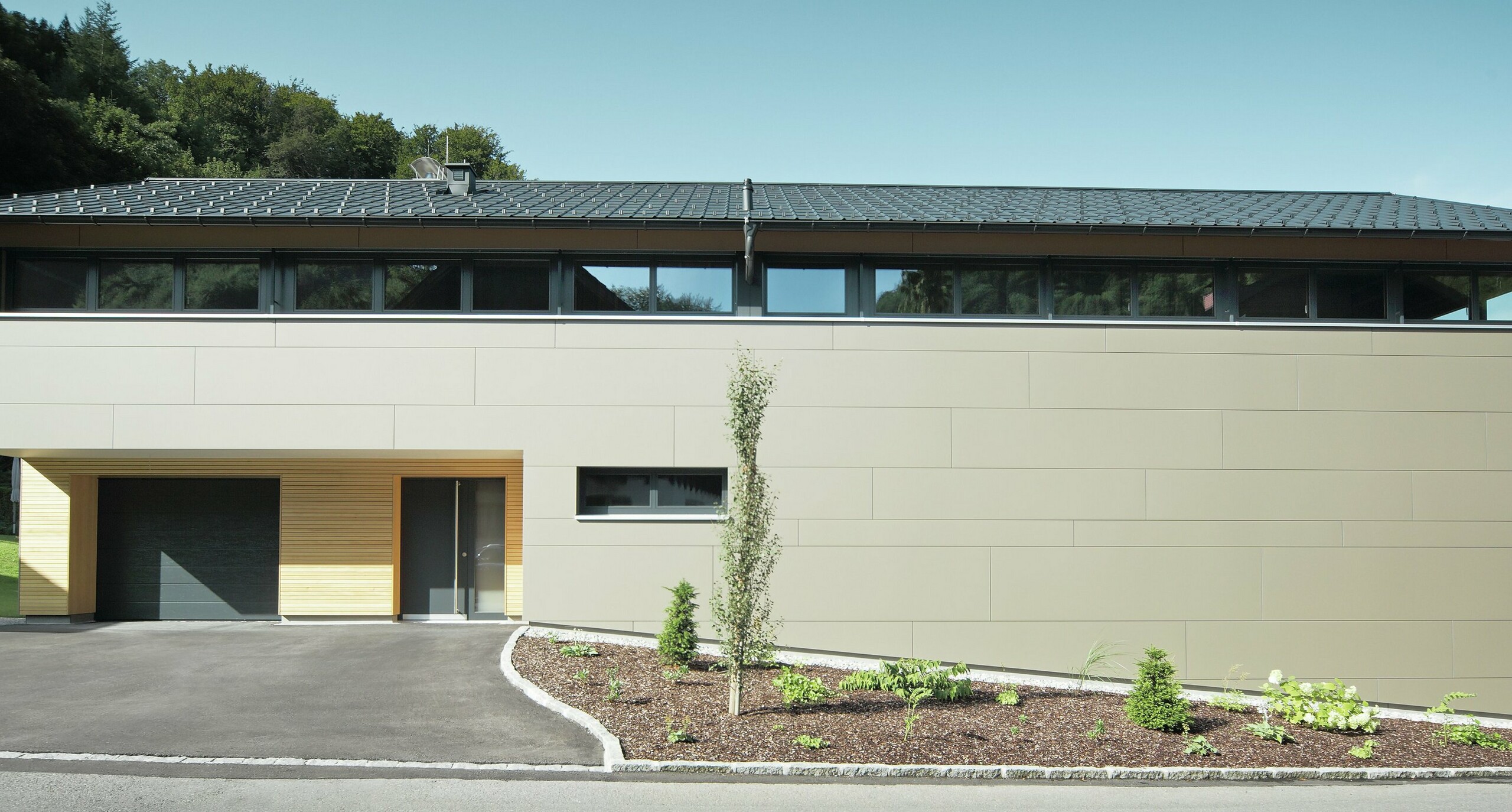 Das Einfamilienhaus in Hohenems präsentiert sich in einer modernen und stilvollen Fassade aus PREFA Aluminium Verbundplatten in Bronzemetallic, mit einem darüberliegenden Dach aus PREFA Dachplatten in P.10 Anthrazit. Die klare Linienführung und das durchdachte Farbkonzept des Designs integrieren das Gebäude nahtlos in die natürliche Umgebung. Die Gestaltung zeigt die Kombination aus Funktionalität und Ästhetik, die PREFA Produkte für zeitgemäße Wohnarchitektur bieten.