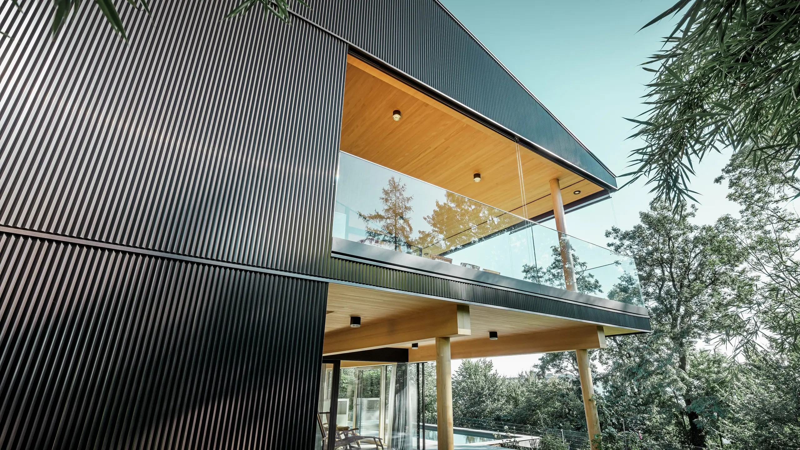 Vue latérale d'une maison individuelle, la façade est revêtue d'un profil triangle PREFA foncé. On aperçoit la terrasse et un grand balcon. Le plafond et la sous-face sont habillés de panneaux de bois.