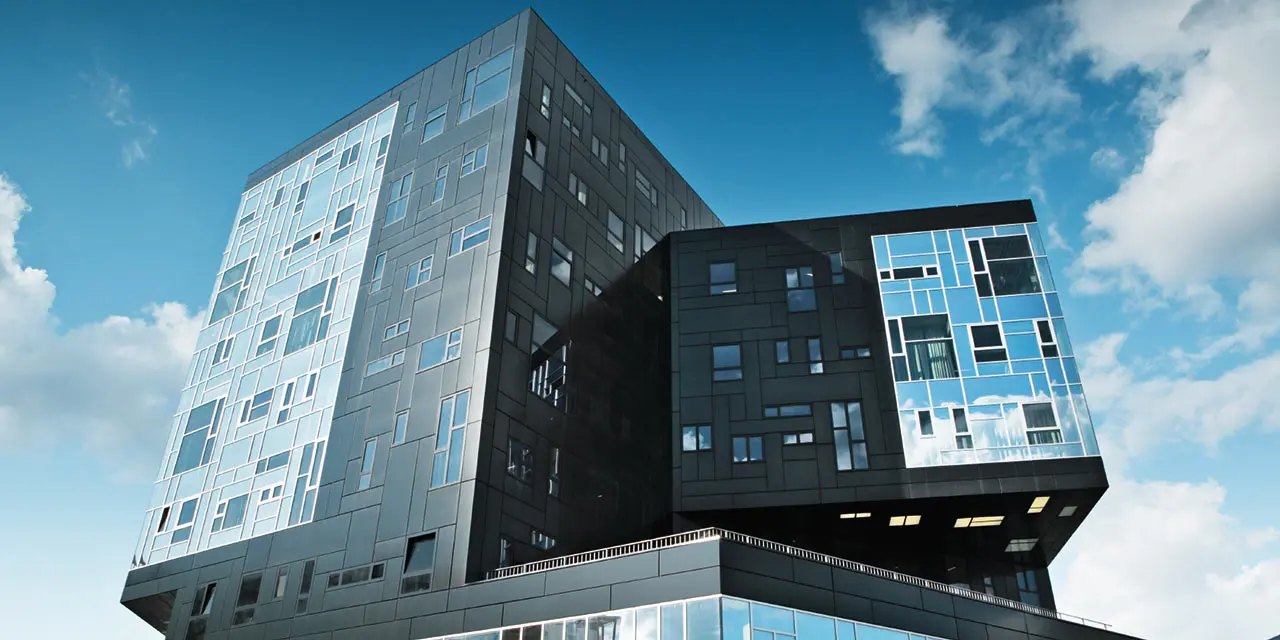 Modernes Universitätsgebäude mit markanter, geometrischer Architektur und überhängenden Baukörpern. Die Fassade besteht aus dunklen und hellen Aluminium-Verbundplatten in unterschiedlichen Grautönen mit unregelmäßig angeordneten Fenstern. Das Gebäude steht unter blauem Himmel mit vereinzelten Wolken und spiegelt das Licht in verschiedenen Facetten wider.