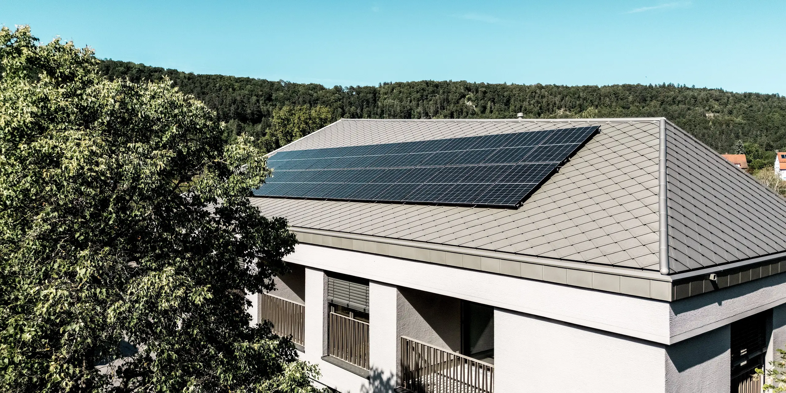 Die Sparkasse Blaubeuren präsentiert sich mit einem eleganten Walmdach aus PREFA Dachrauten 44 × 44 in Bronze. Die großflächige Photovoltaikanlage setzt einen modernen Akzent auf der gleichmäßigen Rautenstruktur. Umgeben von grünen Baumwipfeln und bewaldeten Hügeln überzeugen die leichten, langlebigen und witterungsbeständigen Aluminiumrauten durch zeitlose Ästhetik und hohe Funktionalität.