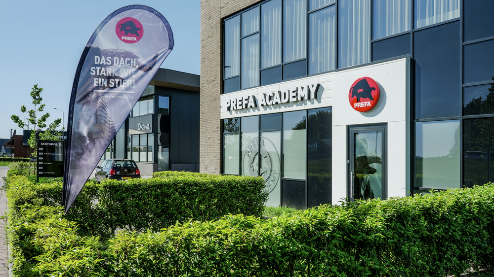 Außenansicht der PREFA Schulungszentrum in Nijkerk mit Werbefahne „Stark wie ein Stier“ und PREFA Logo über der Eingangstür.