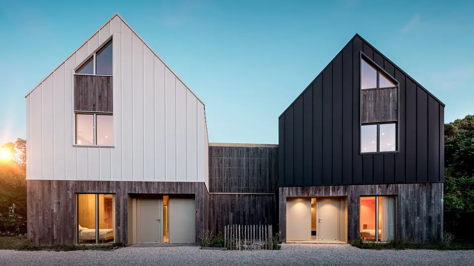 La façade du complexe résidentiel Le Rhune présente deux bâtiments décalés, à l'architecture identique, qui se distinguent par leurs contrastes de blanc et de noir. La pureté des lignes des toitures à deux pans et le revêtement continu en aluminium PREFALZ confèrent aux bâtiments une allure sculpturale et mettent en valeur la précision des joints debout. De chaleureux jeux de lumière, filtrés par les larges fenêtres, créent un contraste saisissant avec l'architecture minimaliste. L'aluminium PREFALZ, résistant aux intempéries et durable, assure la cohésion de la toiture et de la façade, formant une enveloppe de bâtiment moderne et harmonieuse, d'une grande qualité esthétique.