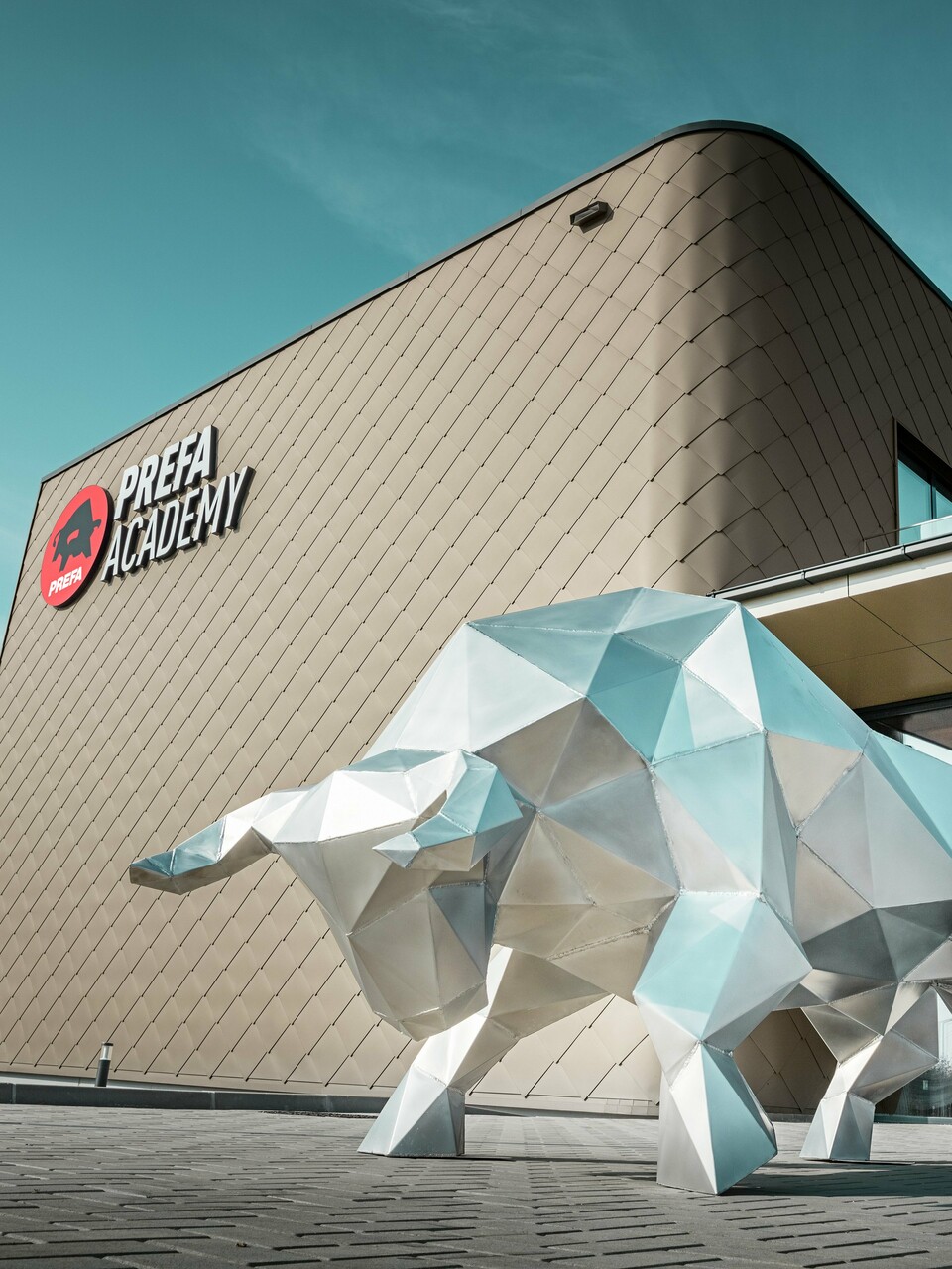 Une imposante sculpture de taureau en aluminium brillant de 2 mm caractérise la zone d'entrée saisissante du nouveau site PREFA à Törökbálint. La façade moderne avec des losanges de façade PREFA 44 × 44 en bronze confère au bâtiment nouvellement construit une enveloppe extérieure élégante, résistante aux intempéries et durable. Les portes coulissantes en verre créent une atmosphère lumineuse dès l'entrée dans le bâtiment. Le logo PREFA Academy, clairement visible sur la façade, souligne le caractère fort du bâtiment de l'entreprise.