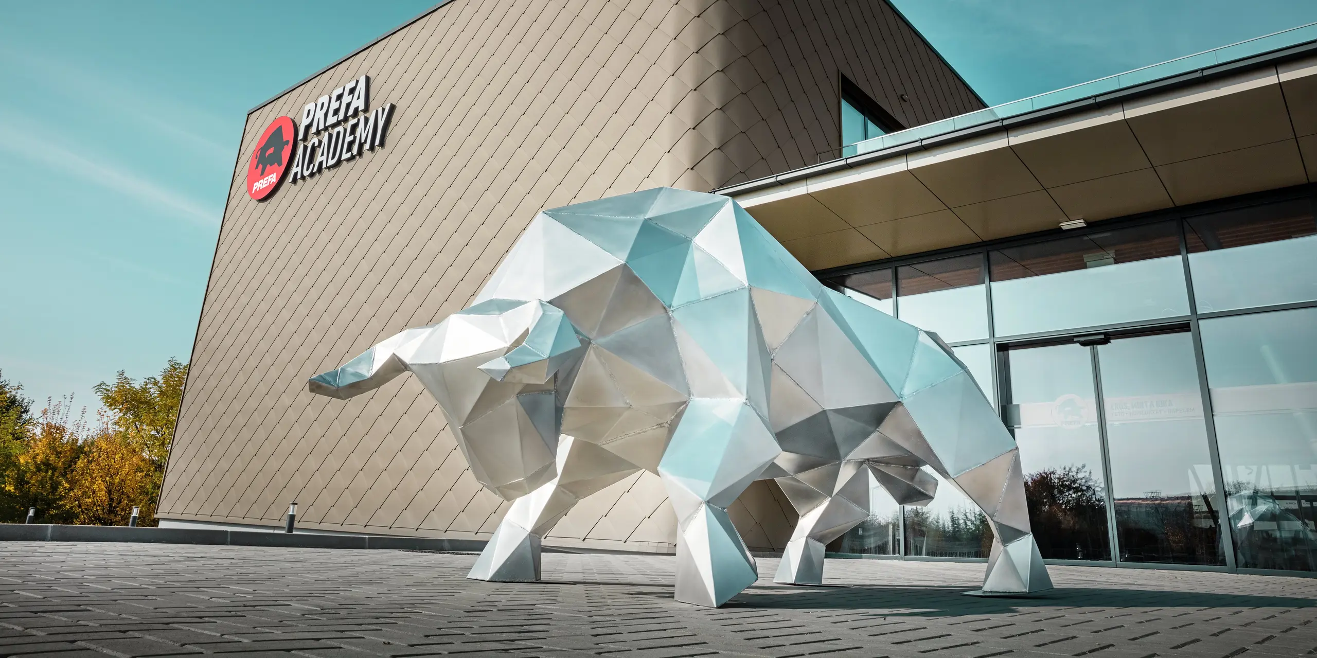 Une imposante sculpture de taureau en aluminium brillant de 2 mm caractérise la zone d'entrée saisissante du nouveau site PREFA à Törökbálint. La façade moderne avec des losanges de façade PREFA 44 × 44 en bronze confère au bâtiment nouvellement construit une enveloppe extérieure élégante, résistante aux intempéries et durable. Les portes coulissantes en verre créent une atmosphère lumineuse dès l'entrée dans le bâtiment. Le logo PREFA Academy, clairement visible sur la façade, souligne le caractère fort du bâtiment de l'entreprise.