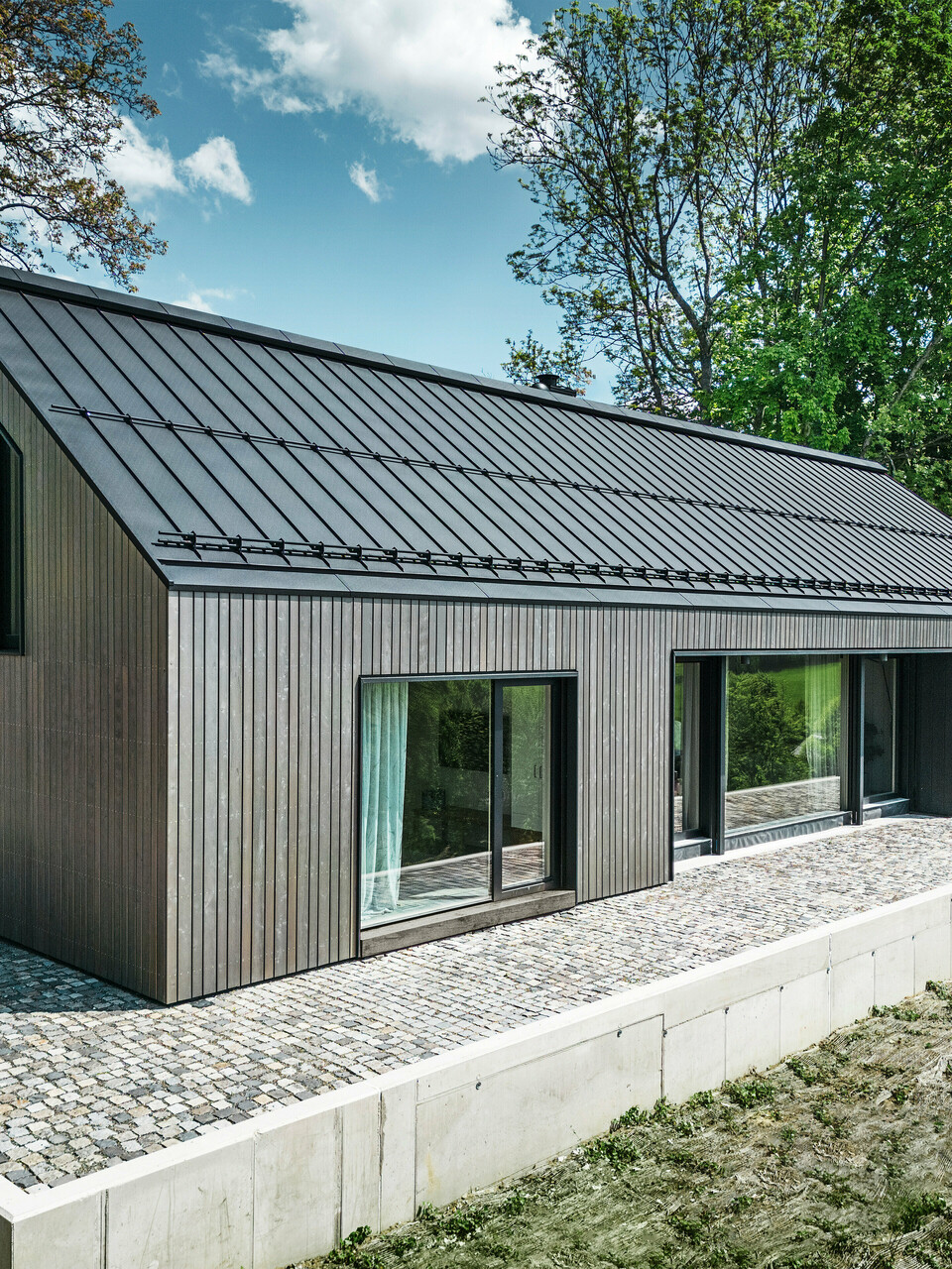 Vorderseite eines Holzmassivhauses in Kuncice, CZ. Das langlebige Aluminiumdach besteht aus PREFALZ in elegantem Schwarz. Das Haus verfügt über großzügige Fensterflächen und einen Whirlpool im Außenbereich.