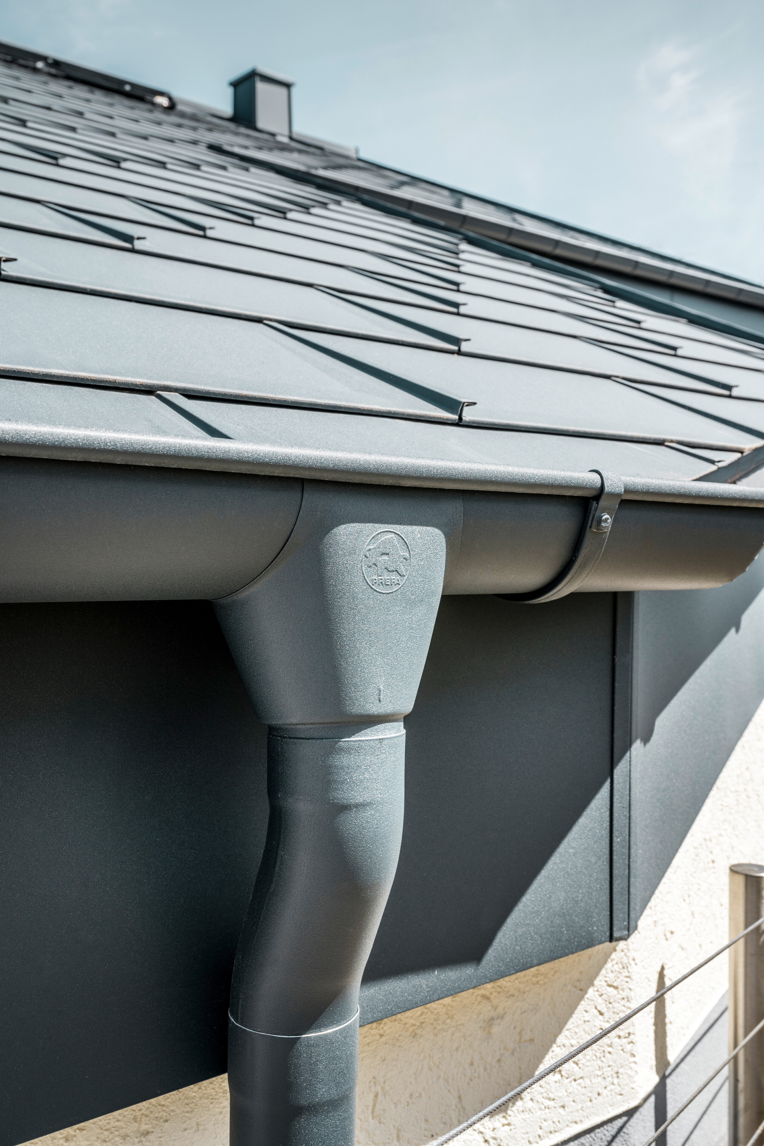 Nahaufnahme eines Hausdaches mit PREFA Dachplatte R.16 in Anthrazit sowie detailreicher Ansicht der farblich abgestimmten PREFA Dachrinne und Fallrohr aus Aluminium bei sonnigem Wetter.