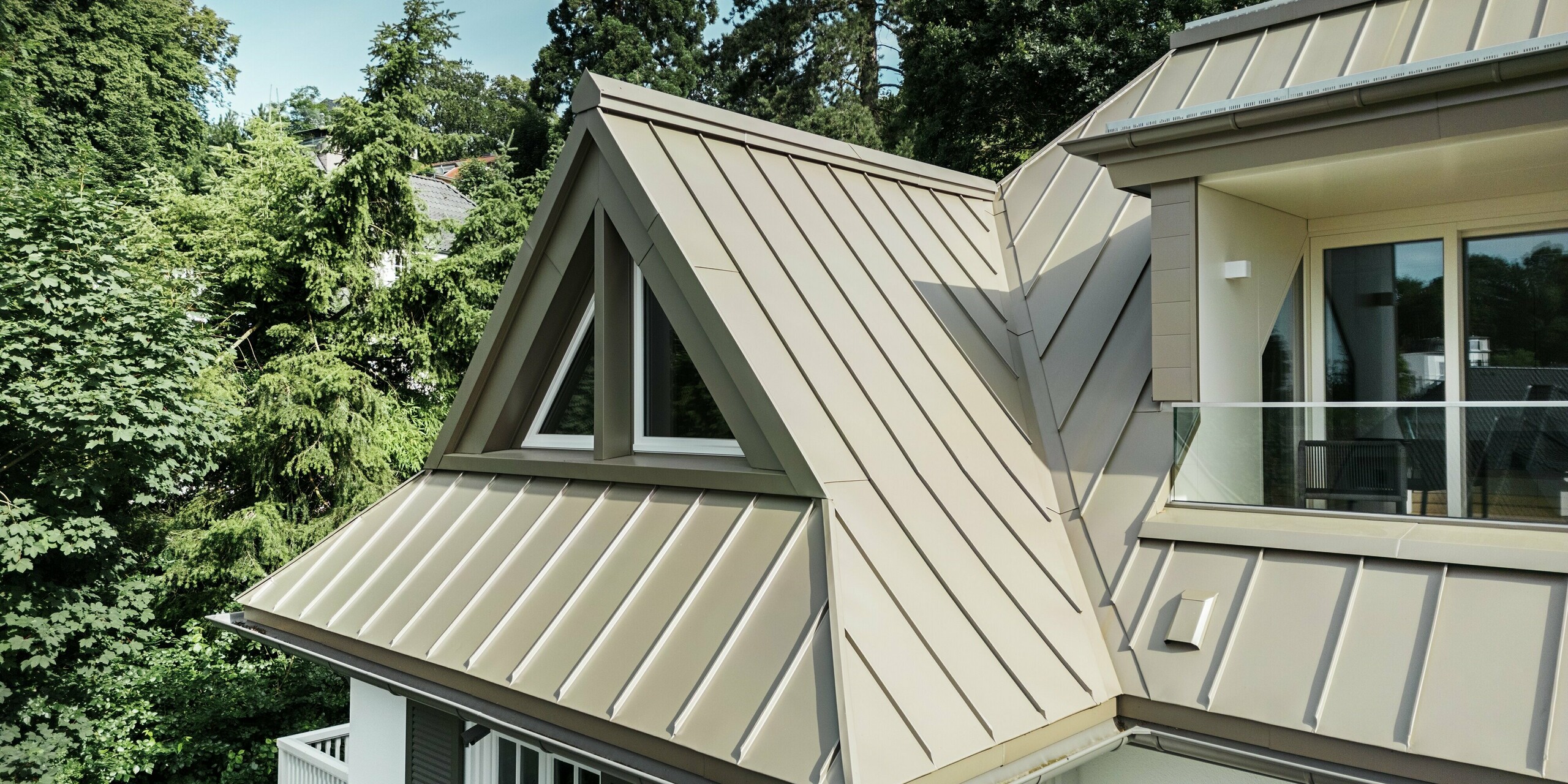 Diese Ansicht zeigt die Dachfenster der Villa Blankenese mit ihren markanten Gauben, die mit PREFALZ in Bronze verkleidet sind. Am rechten Bildrand ist ein eleganter Balkonbereich zu erkennen. Die präzise Stehfalzdeckung aus Aluminium betont die klare Architektur des Gebäudes und sorgt für dauerhaften Schutz vor Witterungseinflüssen. Die bronzene Oberfläche reflektiert das Licht edel und verleiht dem Gebäude eine moderne, hochwertige Optik. PREFA Aluminiumprodukte sind korrosionsbeständig, leicht und wartungsfrei – ideal für langlebige Dächer und Fassaden in anspruchsvollen Bauprojekten.