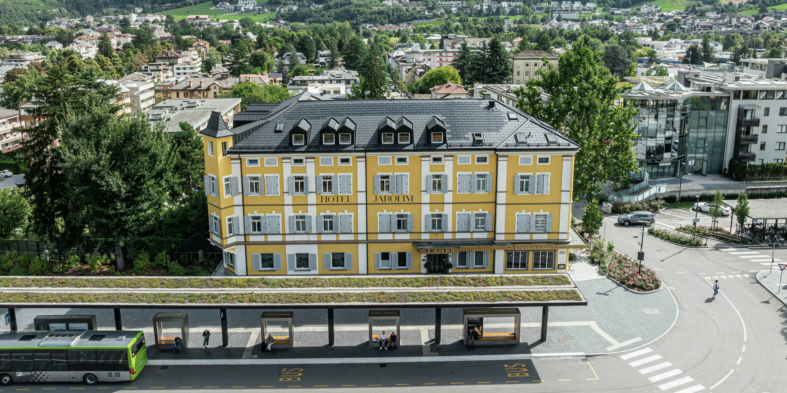 Vue d'en haut de l'hôtel Jarolim rénové à la gare de Bressanone avec un toit composé de tuiles solaires PREFA et de R.16. Le toit robuste en aluminium impressionne par sa construction durable, résistante aux intempéries et légère, idéale pour les bâtiments classés. Les modules photovoltaïques harmonieusement intégrés assurent une production d'énergie durable sans compromettre l'esthétique historique. La tour impressionnante a été conçue avec des tuiles PREFA robustes.