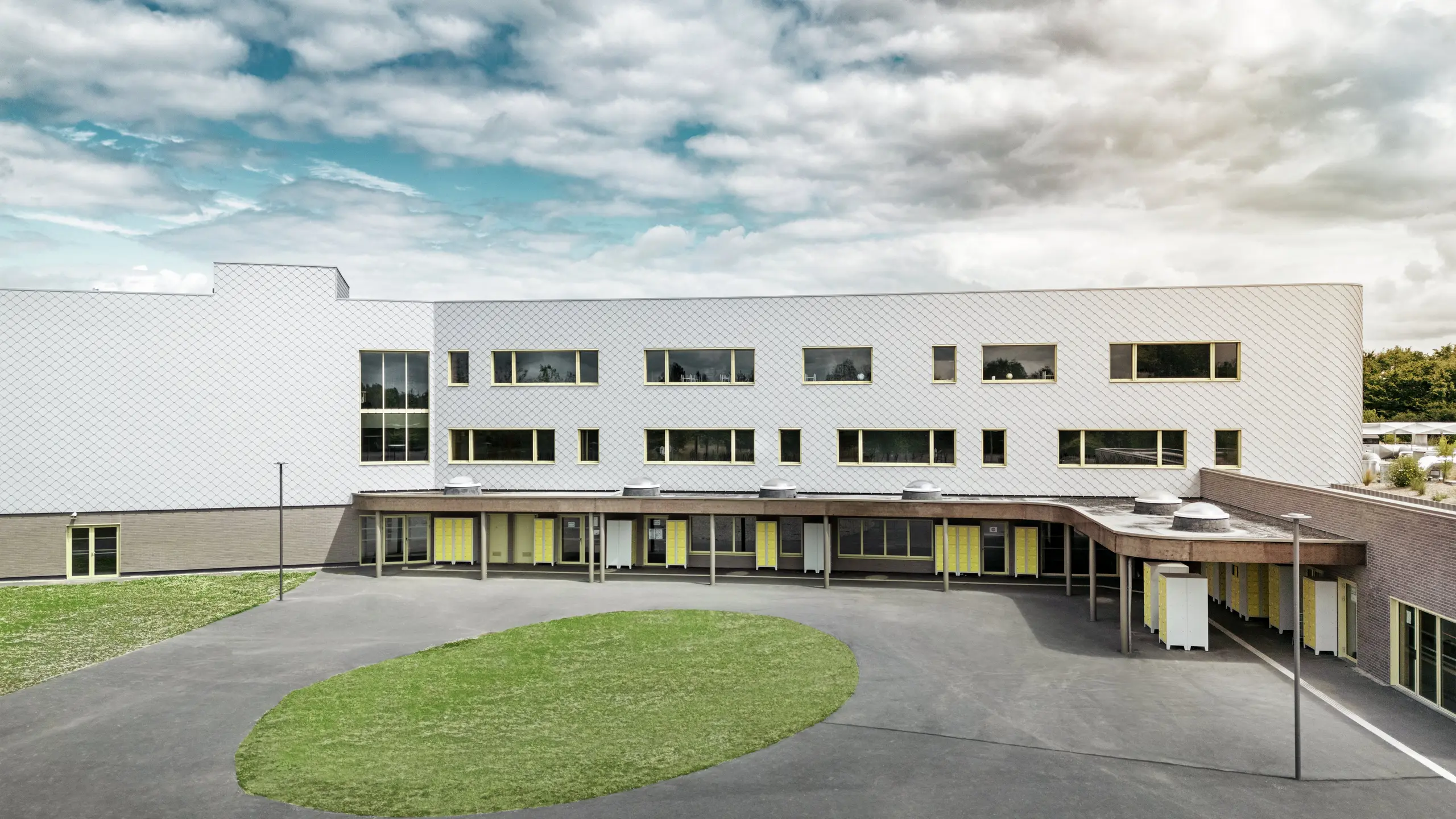 Vue arrière du collège Léontine Dolivet, avec sa grande cour de récréation et ses lignes architecturales épurées. La façade est composée de losanges de façade PREFA argent métallisé, lui conférant un aspect moderne et élégant. Plusieurs rangées de fenêtres et de portes jaunes rythment la structure, tandis que la cour pavée, agrémentée d'un espace vert central, offre un lieu de détente agréable. La façade en aluminium, à la fois résistante et esthétique, allie légèreté, durabilité et design – un atout fonctionnel et esthétique indéniable à Cesson-Sévigné, près de Rennes.