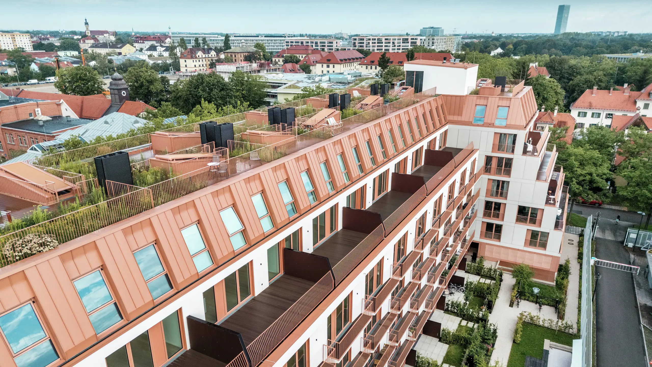 Luftaufnahme des Wohnprojekts Van B in München mit einem markanten Aluminiumdach von PREFA in der Sonderfarbe IGP Orange HDP. Großzügige Balkone, begrünte Dachgärten und eine präzise Fassadengestaltung unterstreichen das durchdachte Architekturkonzept. Ideal für nachhaltige Stadtentwicklung und urbanes Wohnen mit hohem Anspruch an Lebensqualität und Ästhetik.