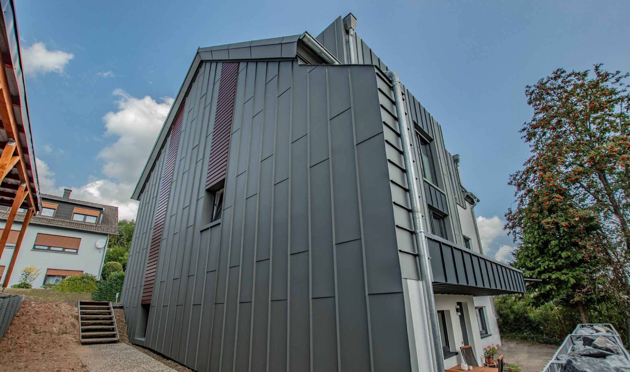 Fassadengestaltung mit vertikaler Winkelstehfalzfassade in hellgrau und Fassadenpaneelen, horizontal verlegt, in Holz dunkel. Die gesamte Fassade wurde aus Aluminium von PREFA gefertigt.