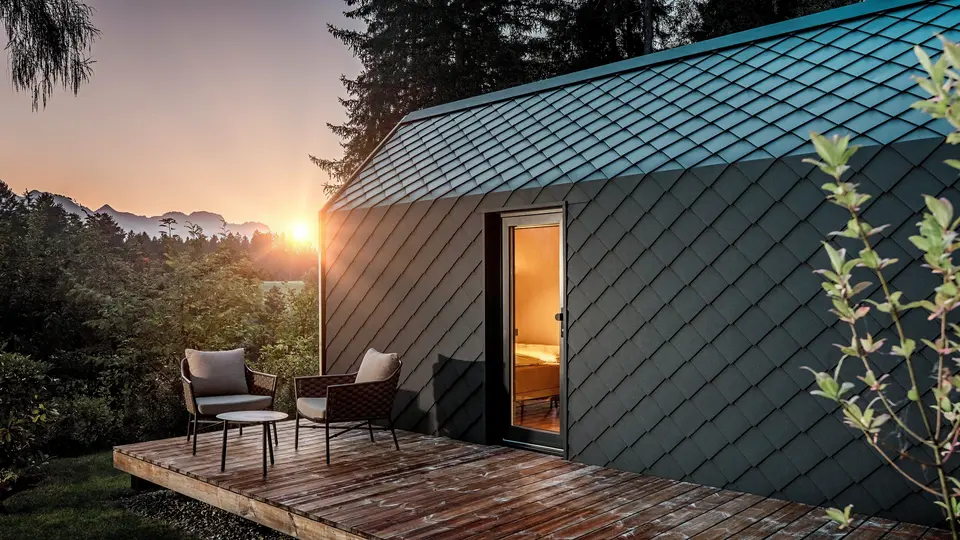 Au coucher du soleil, une minuscule maison minimaliste nichée dans le Secret Koi Garden, sur les rives du lac Natterer, se dévoile. Sa terrasse en bois offre une vue imprenable sur la forêt. Toit et façade sont revêtus de petits losanges en aluminium PREFA P.10 anthracite, dont la surface texturée reflète avec élégance la douce lumière. Durable et résistant aux intempéries, l'aluminium PREFA protège efficacement la structure et crée un contraste saisissant avec le bois, la nature environnante et la lumière tamisée.