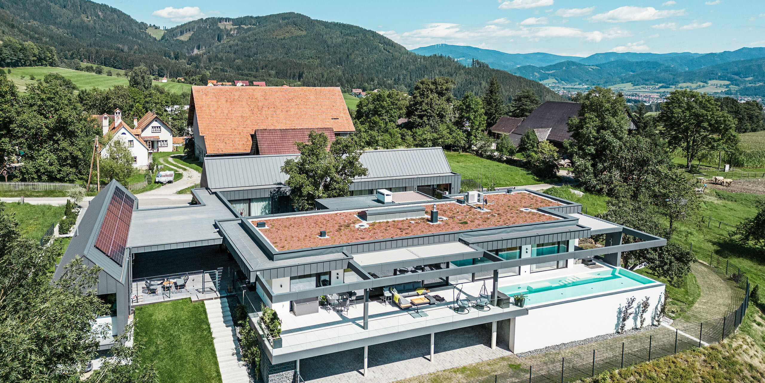 Pohled z dronu na impozantní rodinný dům s částmi budovy využívanými pro zemědělské účely, terasou a bazénem v rakouském Pogier. Tmavě šedé budovy rámuje úchvatná horská krajina. Nemovitost je prezentována střešním a fasádním obkladem PREFALZ v tmavě šedé barvě P.10, který zdůrazňuje jasné, moderní linie. Terasa a bazén nabízí luxusní venkovní obývací prostor s panoramatickým výhledem na okolní přírodu. Kombinace moderních stavebních prvků z PREFA a idylického umístění vytváří dokonalou symbiózu současného designu a přírodních krás.
