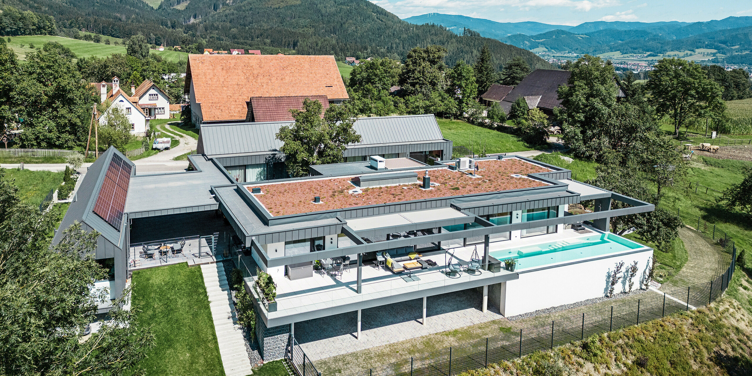 Drohnenaufnahme eines imposanten Einfamilienhauses mit landwirtschaftlich genutzten Gebäudeteilen, Terrasse und Swimmingpool in Pogier, Österreich. Umrahmt werden die dunkelgrauen Gebäude von einer atemberaubenden Berglandschaft. Das Objekt präsentiert sich mit einer PREFALZ Dach- und Fassadenbekleidung in dunkelgrau P.10, die die klaren, modernen Linien unterstreicht. Die Terrasse und der Swimmingpool bieten einen luxuriösen Außenwohnbereich mit Panoramablick auf die umliegende Landschaft. Die Kombination aus den modernen Bauelementen von PREFA und der idyllischen Lage schafft eine perfekte Symbiose aus zeitgemäßem Design und natürlicher Schönheit.