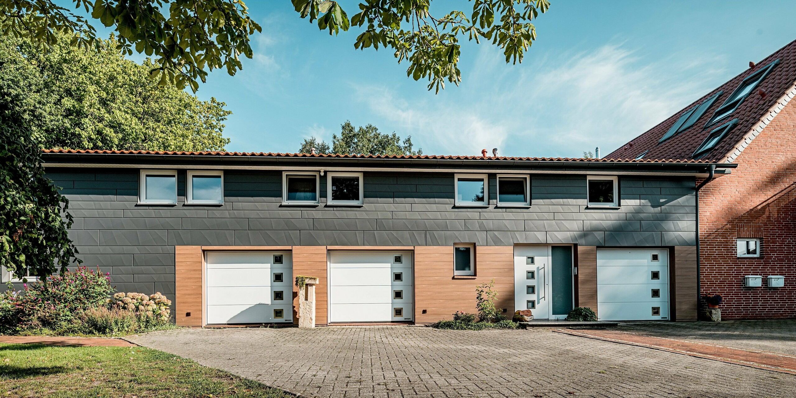 Gesamtansicht eines sanierten Einfamilienhauses mit moderner PREFA Aluminiumfassade in klarer, horizontaler Linienführung. Die Fassade kombiniert PREFA Siding.X in P.10 Dunkelgrau im oberen Bereich mit PREFA Siding in Walnussbraun im Sockel- und Eingangsbereich und schafft so einen spannenden Kontrast aus kühler Eleganz und warmer Holzoptik. Die markanten Kantungen der Siding.X-Paneele erzeugen lebendige Licht- und Schattenspiele, während die Holzoptik gezielt Akzente setzt. Weiße Garagentore und präzise Fensteröffnungen unterstreichen das moderne Erscheinungsbild. Die langlebigen, witterungsbeständigen und pflegeleichten PREFA Aluminiumpaneele machen die Fassadensanierung nachhaltig, hochwertig und architektonisch ausdrucksstark.