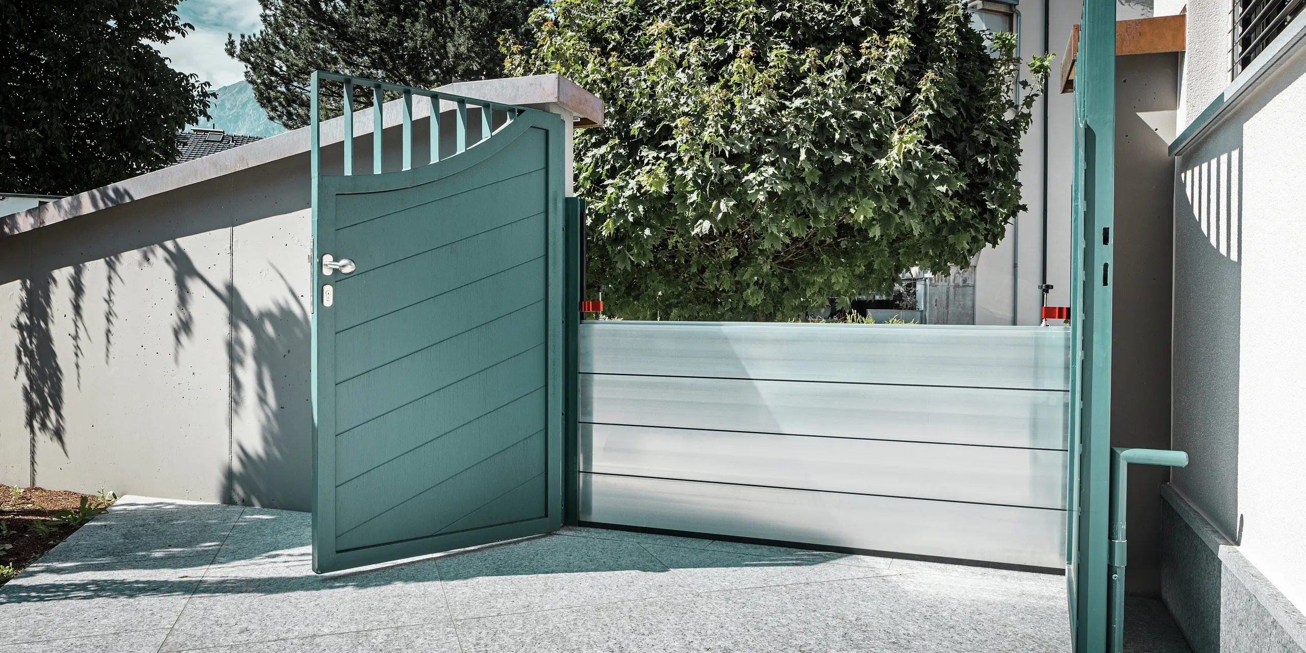 Offenes Gartentor mit integriertem PREFA Hochwasserschutzsystem vor einem Einfamilienhaus in Schwaz, Tirol. Die Aluminium Dammbalken sichern den Eingangsbereich zur Hauswand und bieten einen effektiven Objektschutz bei Überflutungen. Das System fügt sich unauffällig in die bestehende Architektur ein und unterstreicht, wie sich funktionale Sicherheit und ästhetisches Design optimal verbinden lassen.