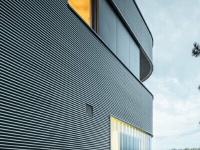 Vue d'ensemble de la façade de la maison de Leonding à Linz, au crépuscule, recouverte avec les profils sinus PREFA dans la teinte anodisé naturel. La lumière se reflète dans la maison.