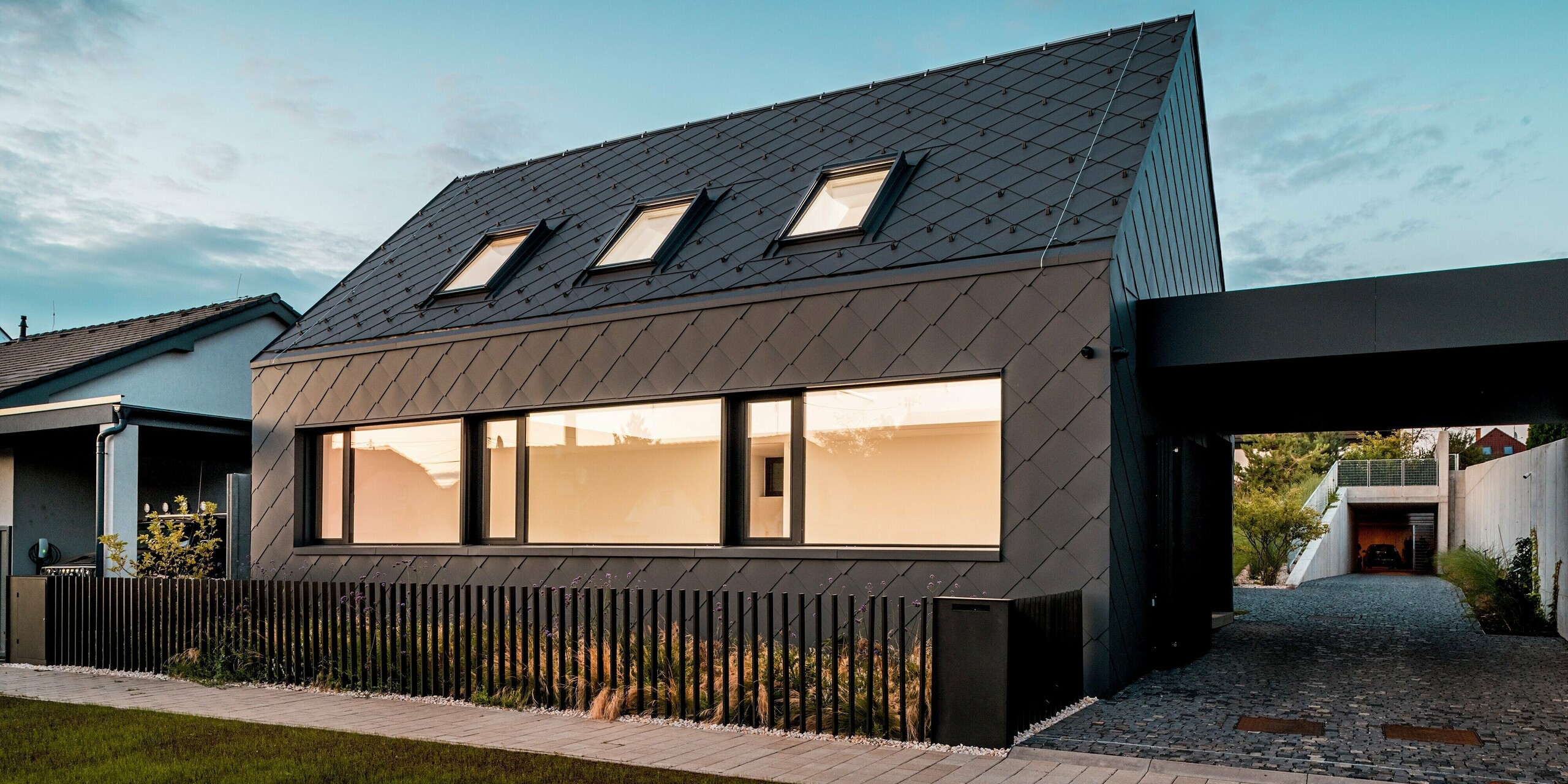 Das Bild zeigt das moderne Einfamilienhaus in Trnava‑Modranka in der Abenddämmerung, komplett verkleidet mit PREFA Dach- und Wandrauten 44 × 44 in P.10 Dunkelgrau. Die beleuchteten Fenster erzeugen einen warmen Kontrast zur dunklen, gleichmäßig strukturierten Aluminiumhülle. Die präzise verlegte Rautenoptik betont die klaren Linien des Baukörpers und unterstreicht die hochwertige, robuste Materialität. Die langlebigen PREFA Aluminiumprodukte bieten optimalen Witterungsschutz und verleihen dem Gebäude ein markantes, zeitloses Erscheinungsbild.