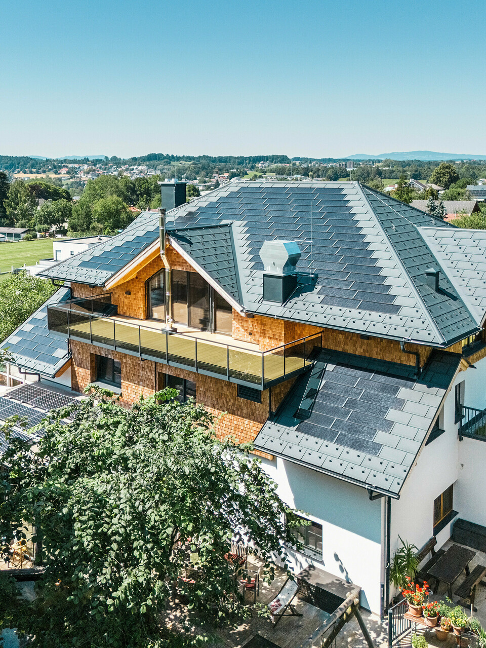 Vue aérienne du Zenz'n Stub'n à Schörfling am Attersee avec un toit récemment rénové en tuiles solaires PREFA et panneaux de toit R.16 en P.10 anthracite. La toiture en tôle de haute qualité allie un design moderne à une production d'énergie durable. Le balcon spacieux et la gouttière harmonieusement intégré soulignent l'aspect élégant du bâtiment.