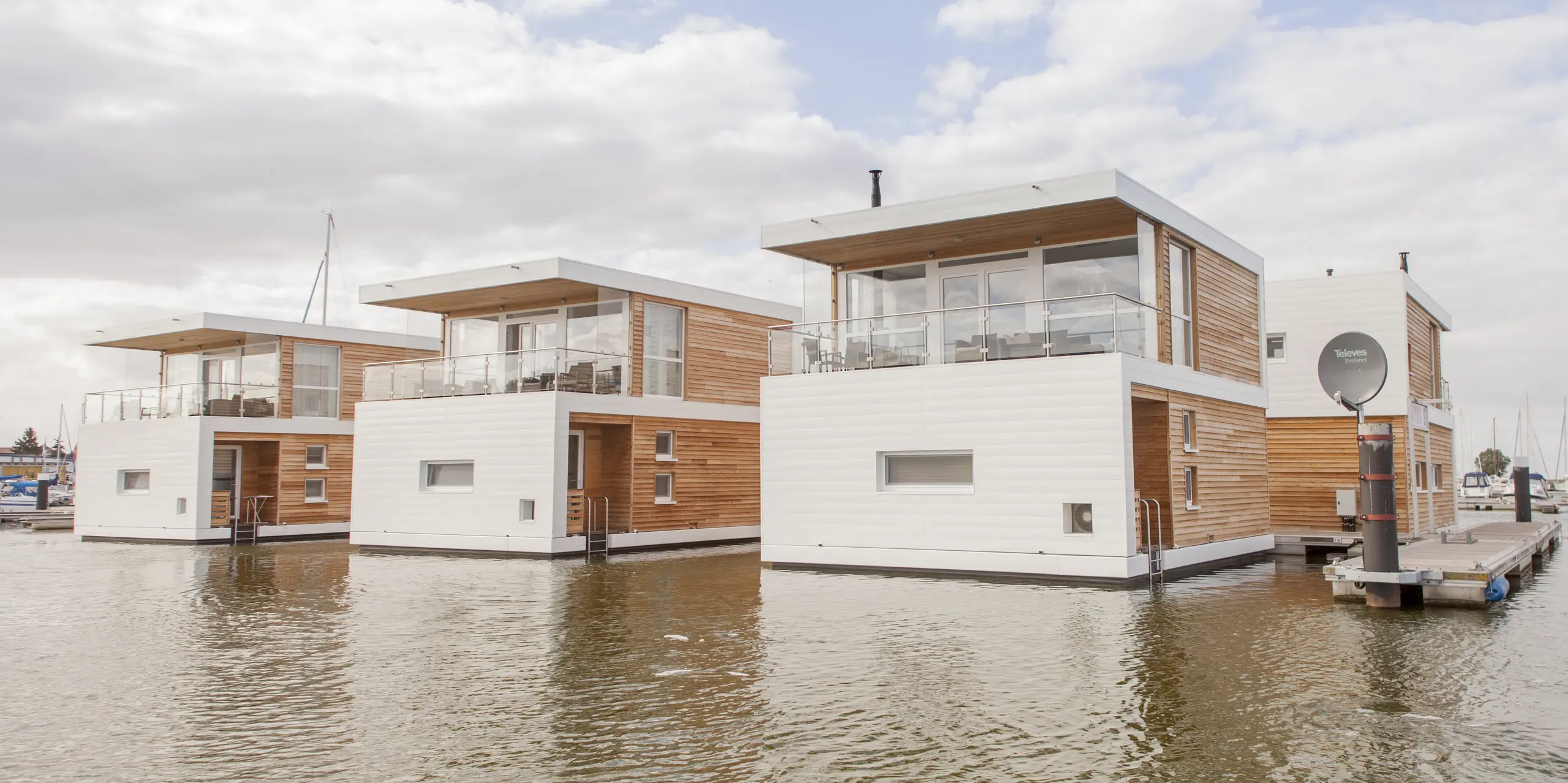 Drei moderne schwimmende Häuser in Stralsund liegen direkt im Wasser des Yachthafens und beeindrucken mit klarer, geradliniger Architektur. Die PREFA Sidings in P.10 Prefaweiß setzen markante Akzente und bilden einen eleganten Kontrast zu den warmen Holzfassaden. Die langlebigen, korrosionsbeständigen Aluminiumfassaden sind ideal für das maritime Umfeld und überzeugen durch ihre Witterungsbeständigkeit sowie Pflegeleichtigkeit.