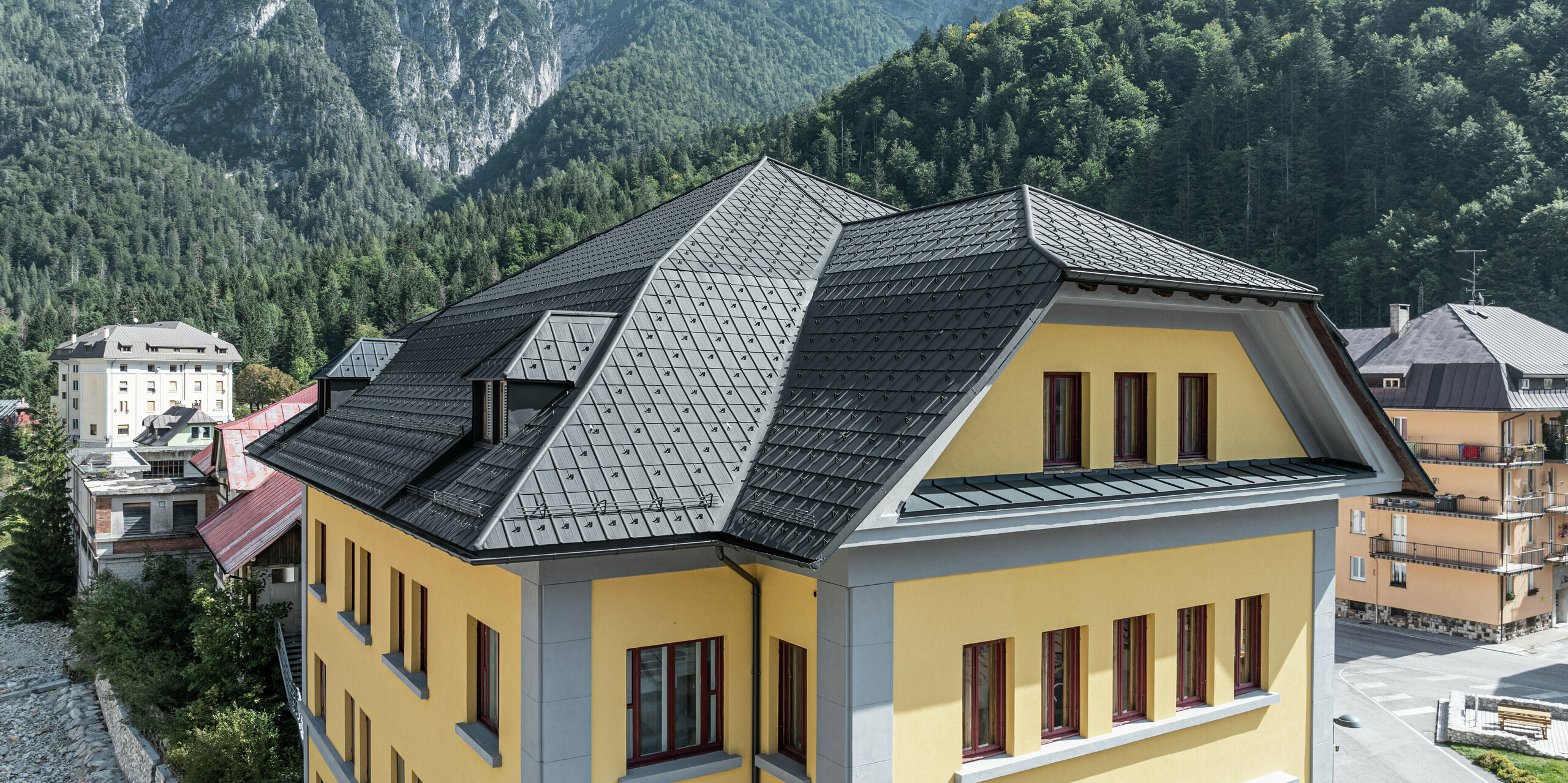 Historisches Museumsgebäude Museo Cave del Predil stilvoll saniert mit robusten PREFA Dachplatten in P.10 Anthrazit. Die moderne PREFA Aluminiumlösung schützt das Gebäude zuverlässig und dauerhaft vor alpinen Witterungseinflüssen. Im Hintergrund die eindrucksvolle Berglandschaft mit bewaldeten Hängen, die die Anforderungen an Robustheit und Witterungsbeständigkeit unterstreicht. Ideale Dachlösung von PREFA für den Einsatz in Regionen mit hohen Schneelasten und anspruchsvollen klimatischen Bedingungen.