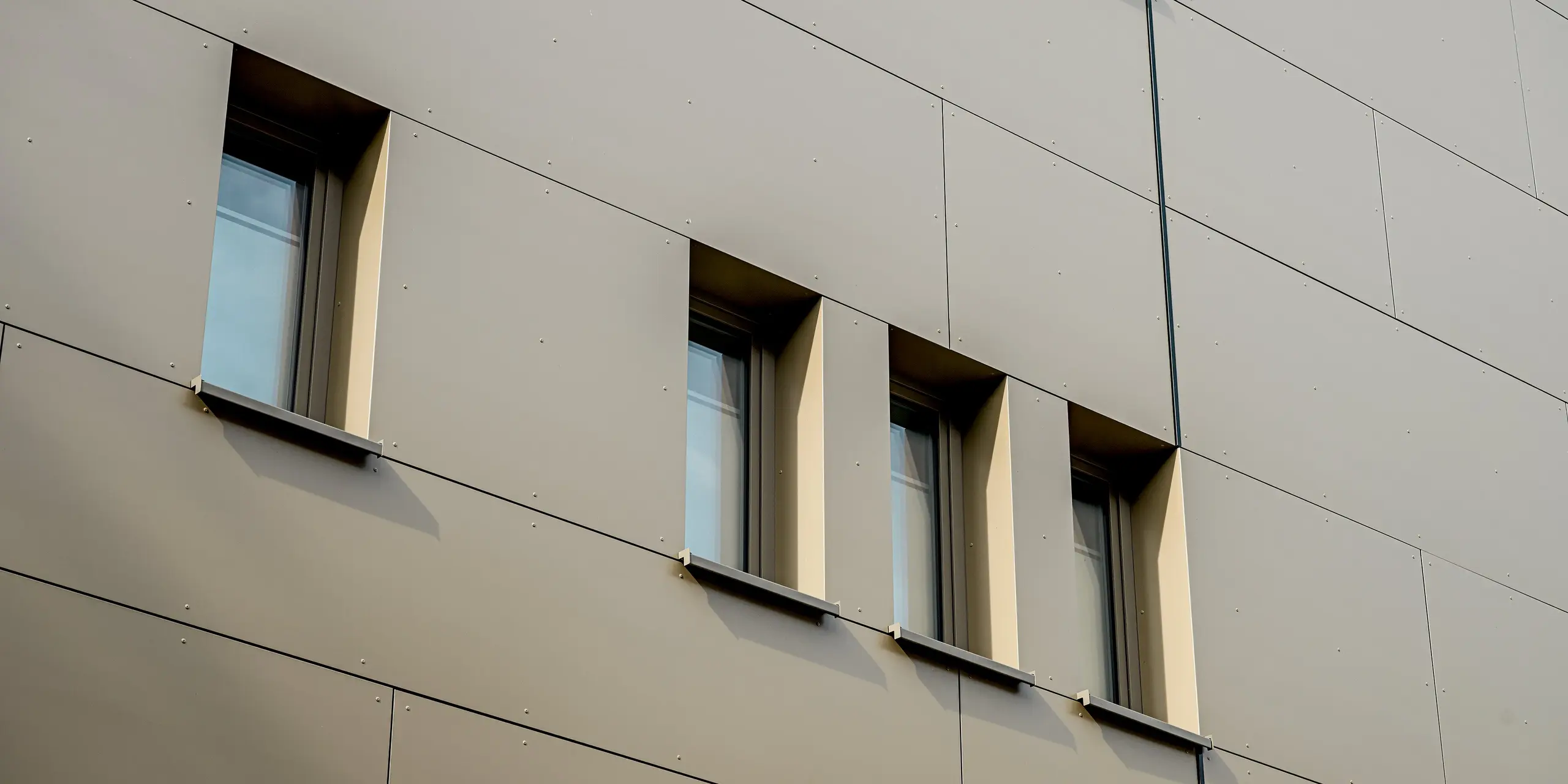 L'image présente un gros plan de la façade du bâtiment Festo à Langenhagen. On y observe d'élégants panneaux composites en aluminium PREFABOND, à la finition bronze clair, qui confèrent à la façade une esthétique moderne et apaisante. Les fenêtres, encastrées dans des niches aux formes spécifiques, soulignent le caractère minimaliste du bâtiment. La disposition précise des fenêtres et la finesse des lignes mettent en valeur la pureté du design. Grâce à la haute résistance et à la durabilité des matériaux, la façade conservera son esthétique et sa fonctionnalité pendant de nombreuses années, ce qui en fait un choix idéal pour une architecture pérenne.