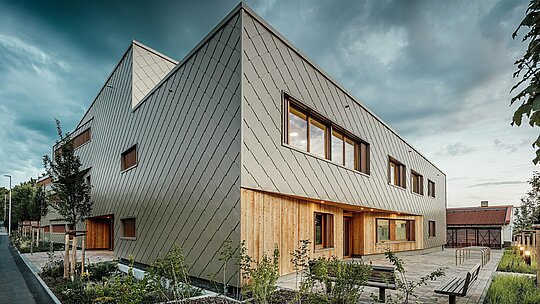 Sous la douce lumière du soir, le bâtiment hybride de la crèche d'Alzenau révèle toute sa dimension architecturale. La façade remarquable, composée de losanges de façade PREFA couleur bronze (44 x 44 cm), confère au bâtiment une enveloppe élégante et résistante aux intempéries, à la structure épurée. Au rez-de-chaussée, un bardage vertical en bois offre un contraste naturel et chaleureux avec l'aluminium et met en valeur l'entrée ainsi que les espaces de détente abrités. Le design minimaliste et moderne s'harmonise avec les espaces extérieurs soignés, où bancs, supports à vélos et jeunes arbres invitent à la flânerie. Cette image illustre parfaitement comment les produits PREFA, d'une grande durabilité, contribuent à la création d'un bâtiment éducatif durable, fonctionnel et accueillant.