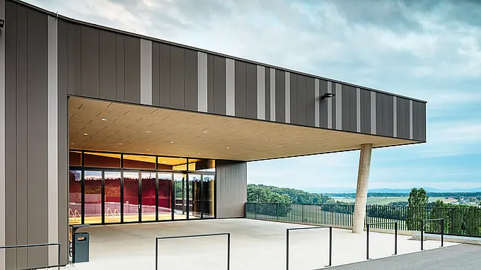 Vue extérieure du centre événementiel ZIB Nikolai à St. Veit, avec une façade moderne en aluminium dans les tons marron et beige-gris. L'entrée est couverte, soutenue par une colonne de béton inclinée, et offre une vue sur le paysage verdoyant environnant et les collines lointaines sous un ciel nuageux.