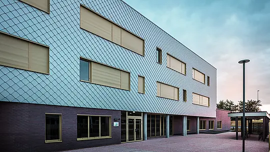 La photo montre l'entrée du Collège Léontine Dolivet, dont la façade moderne est composée de losanges de façade PREFA argent métallisé. Plusieurs fenêtres rectangulaires aux cadres jaunes et aux volets roulants fermés structurent la vue. Au premier plan se trouve une place pavée, bordée d'un espace couvert. La façade apparaît élégante, résistante aux intempéries et durable.