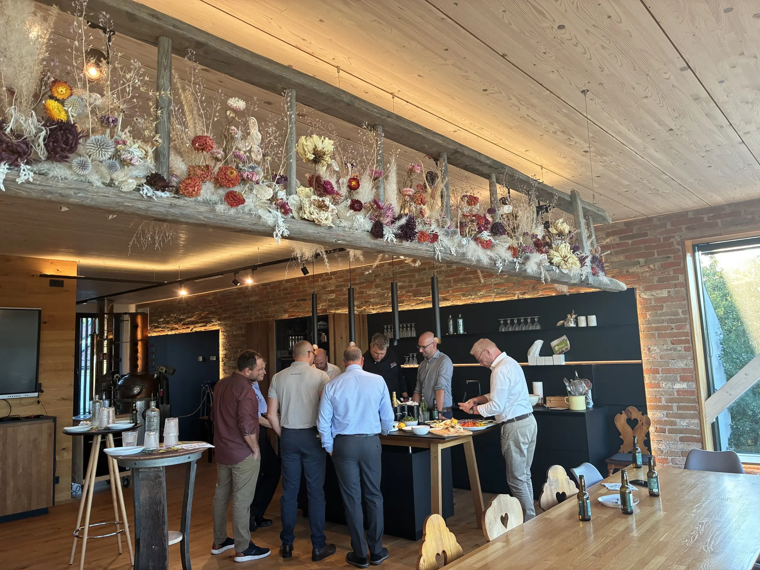 Eine Gruppe von Personen steht während eines gemeinsamen Treffens rund um eine Kücheninsel in einem gemütlichen Raum mit Holz, Ziegelwänden und Blumendekoration.
