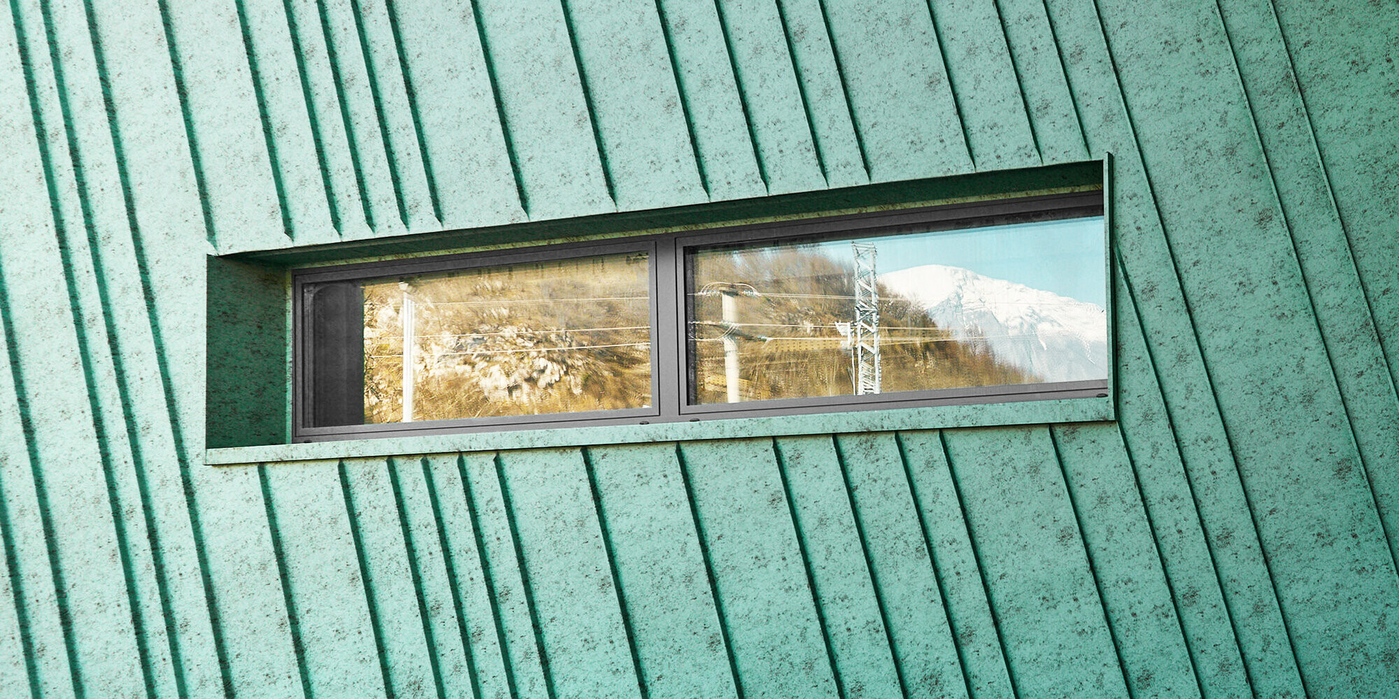 Detail mit leicht schräg verlaufender Fassade mit kleinem, länglichem Fenster, in dem sich Berge und elektrische Leitungen widerspiegeln. Seitenansicht auf Fassade und Glasfront mit Eingang.