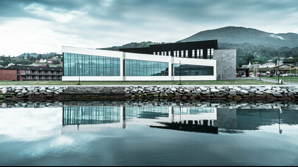 Dans ce cliché, nous voyons les reflets de la façade du centre de santé Hardangerbadet à Oystese dans l'eau. Celle-ci a été recouverte en losange de façade 44x44 en aluminium naturel. On peut apercevoir un bout du Fjord Hardangerfjord dans le fond.