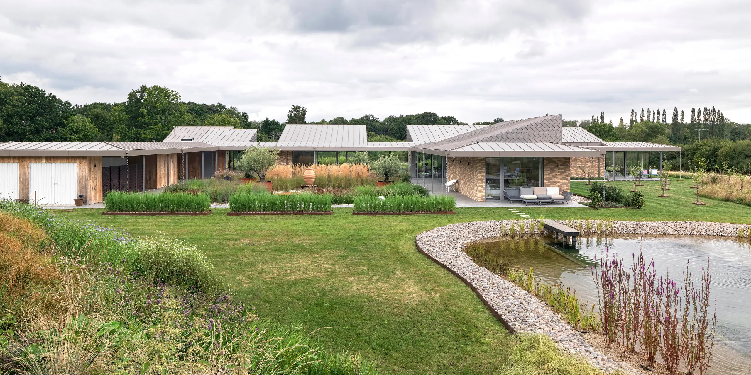 Vue panoramique de cet immeuble résidentiel à l'architecture sophistiquée situé dans le West Sussex, avec sa grande toiture en aluminium PREFALZ couleur P.10 bronze et ses losanges de façade PREFA harmonieusement intégrés à certains endroits de la façade. Ce bâtiment moderne s'étend sur toute la propriété, plat et spacieux, au cœur d'une végétation naturelle et avec un bassin naturel paysager au premier plan. Les robustes produits PREFA s'inscrivent dans le concept de construction durable et garantissent un aspect uniforme et durable.